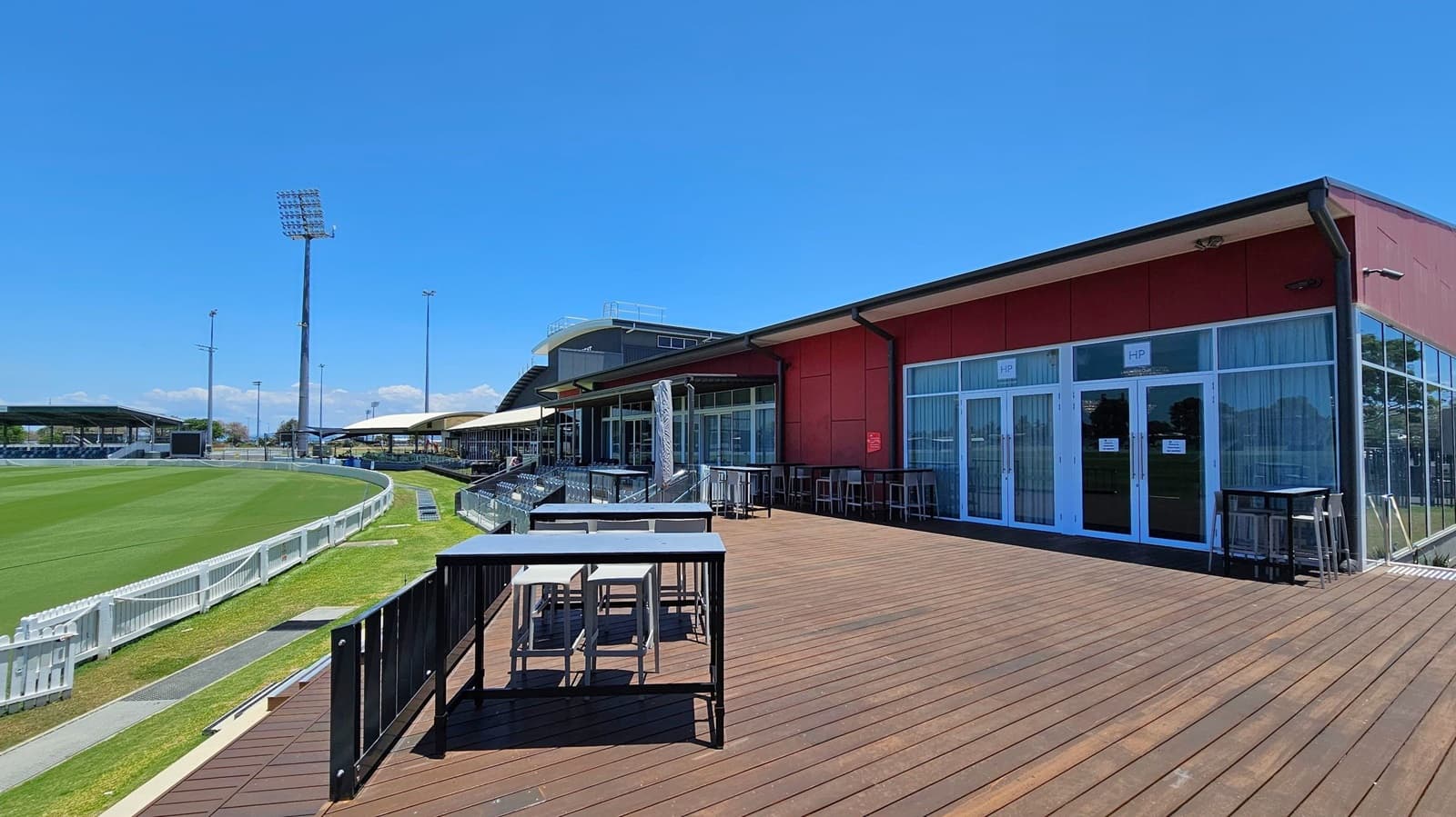 A cricket ground with a well-maintained green field on the left, bordered by a white picket fence. In the background, there are tall floodlights and covered seating areas. On the right, a modern building with large glass windows and doors, featuring a wooden deck with tables and chairs. The deck is spacious, with a clear view of the field.