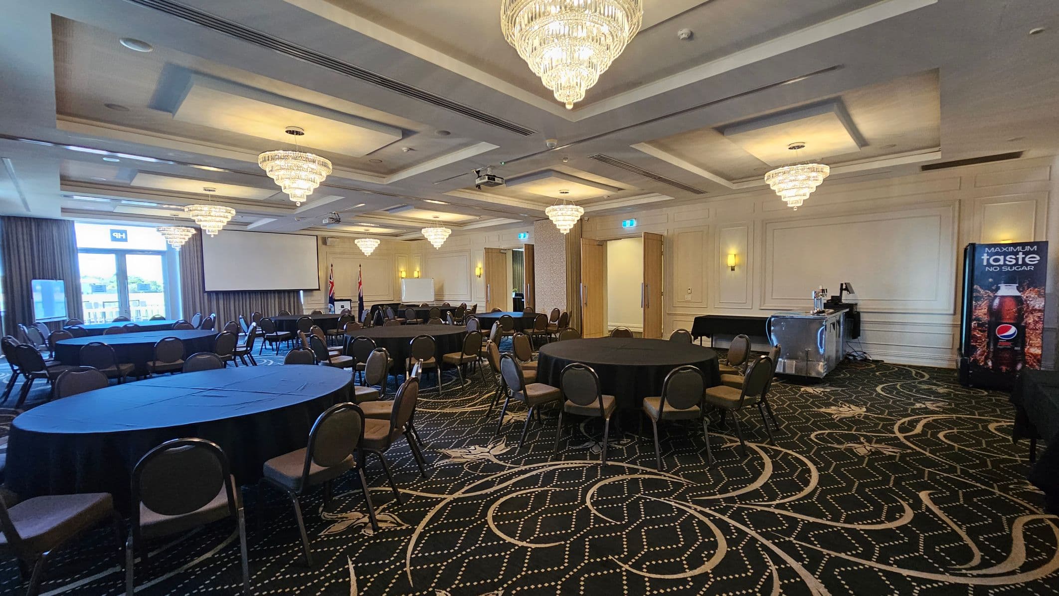 A spacious conference room with round tables covered in black tablecloths and surrounded by chairs. On the left, large windows with curtains allow natural light in. In the centre, a large projection screen is visible. On the right, a drinks station with a Pepsi vending machine is present. The ceiling features multiple ornate chandeliers, and the carpet has a geometric pattern in dark tones. The walls are decorated with moulding and light fixtures.