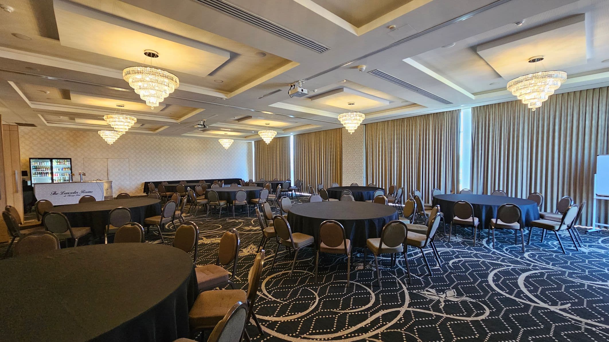A spacious conference room with round tables covered in black tablecloths, surrounded by chairs. On the left, there's a small bar area with a sign reading "The Lavada Room" and a fridge stocked with drinks. The right side features large windows with beige curtains. The ceiling is adorned with multiple crystal chandeliers, and the floor has a patterned carpet in dark tones.