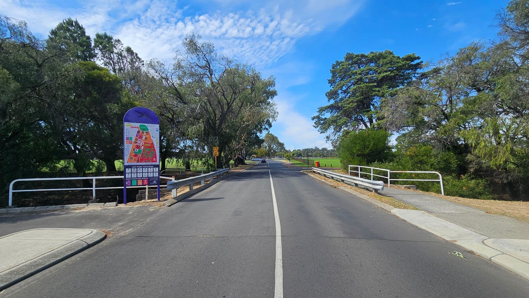 A straight road with a white centre line runs through the middle, flanked by metal railings on both sides. On the left, there's a colourful map sign with trees behind it. On the right, more trees and a red bin are visible. The road leads into a park-like area with more greenery and a few distant cars. The sky is blue with scattered clouds.