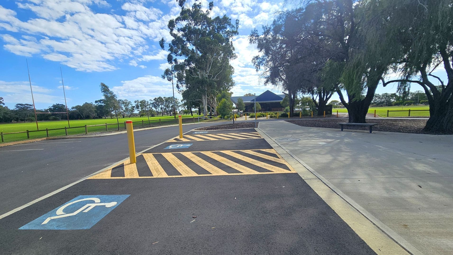 A paved area with a marked disabled parking space on the left, featuring a blue symbol. Yellow and black striped lines are adjacent to the parking space. A yellow bollard stands nearby. On the right, a concrete path runs alongside trees with dense foliage. In the background, a grassy field with rugby posts is visible, along with a building partially obscured by trees. A bench is positioned near the path.