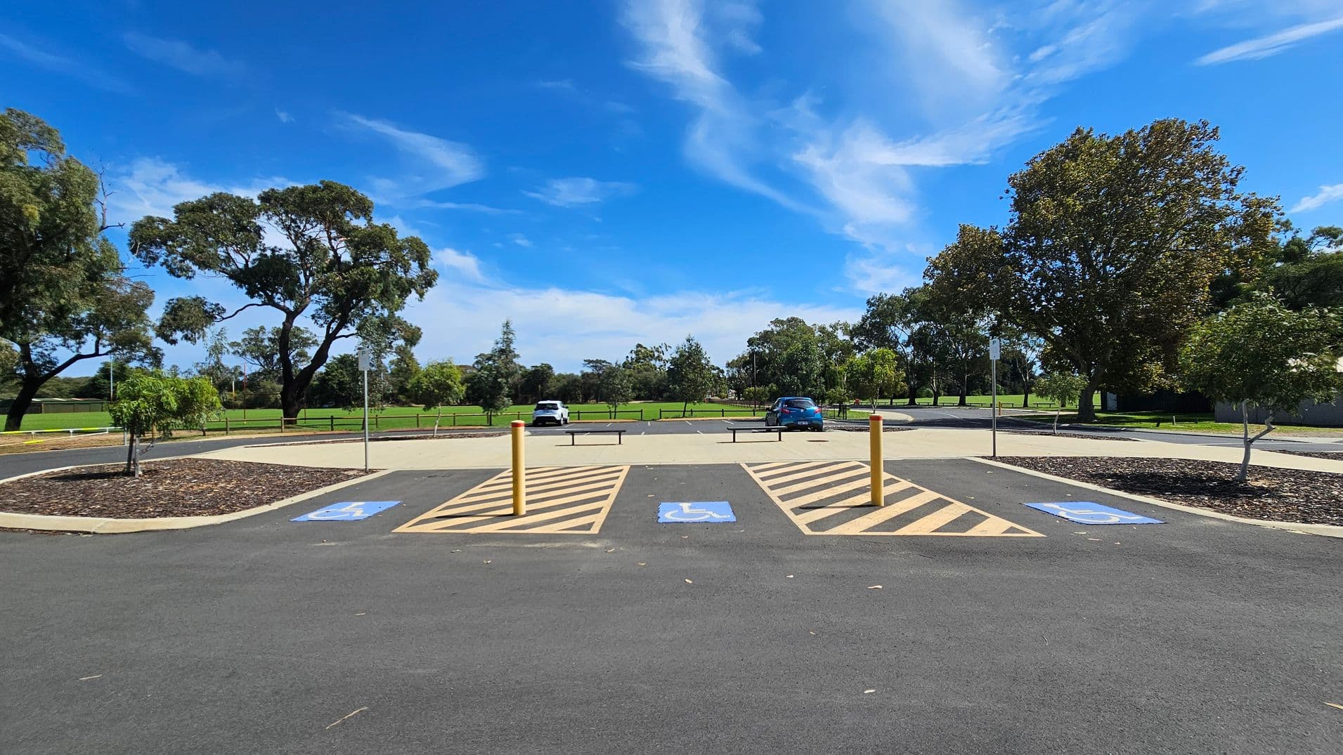 A car park with designated disabled parking spaces marked by blue signs on the asphalt. Yellow bollards separate the spaces. In the background, there are two cars parked, one white and one blue. Surrounding the car park are green grassy areas and several large trees. The sky is clear with some wispy clouds.