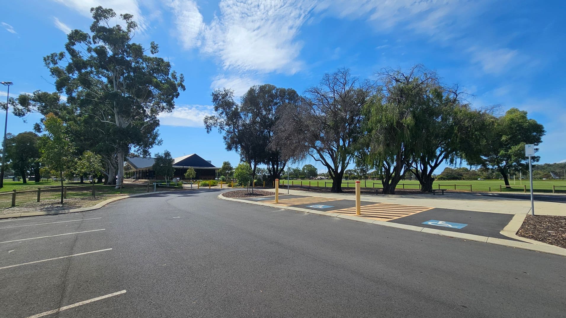 A paved parking area with marked spaces, including a designated disabled parking spot on the right. A row of tall trees lines the background, with a building partially visible to the left. The area is bordered by a grassy field and wooden fencing. The sky is clear with some clouds.