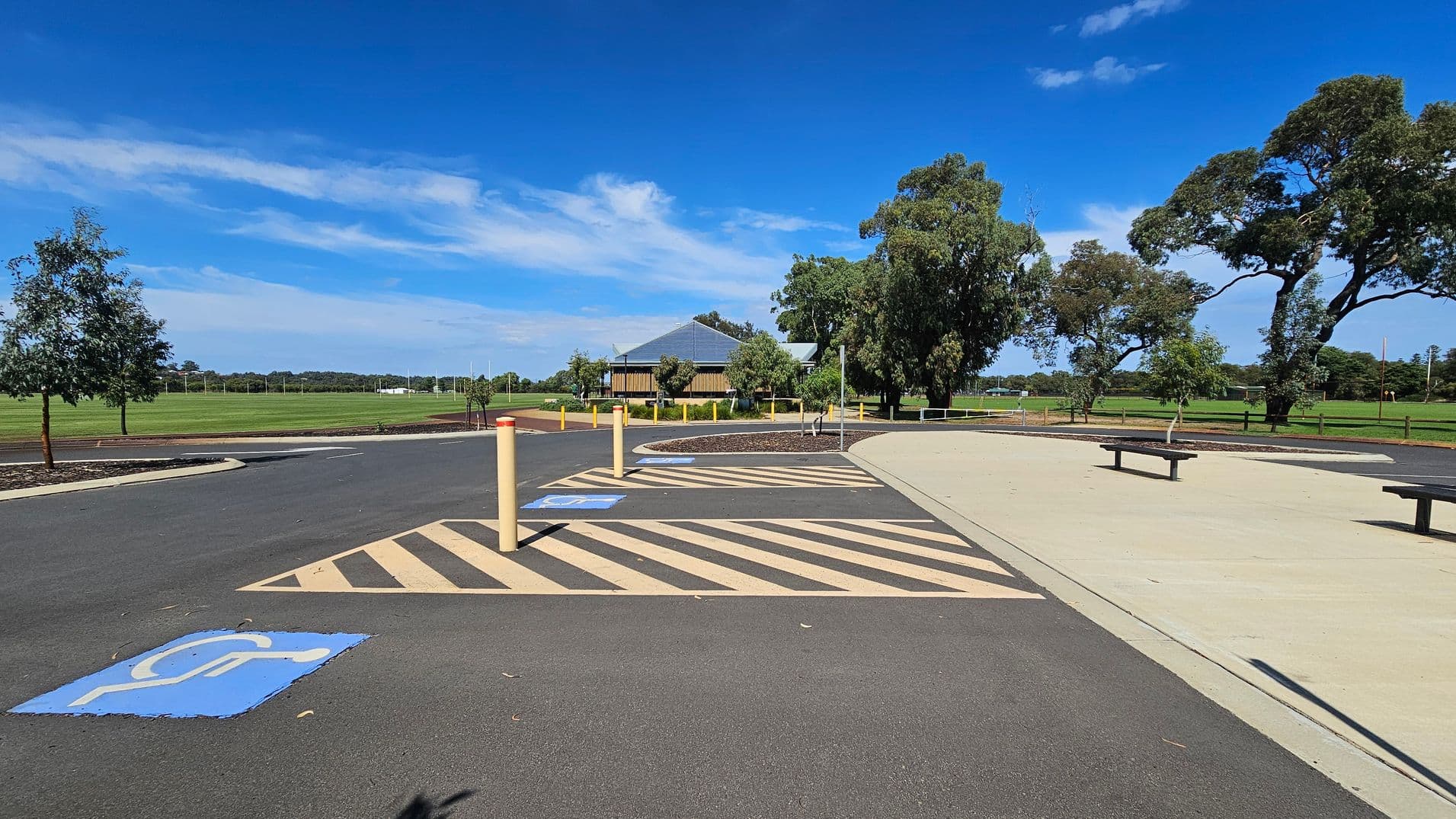 A paved parking area with marked disabled parking spaces and yellow-striped zones. In the centre, a small building with a sloped roof is surrounded by trees. To the right, a concrete path with benches runs parallel to the parking area. The background features a large grassy field with more trees and a fence. The sky is clear with some clouds.