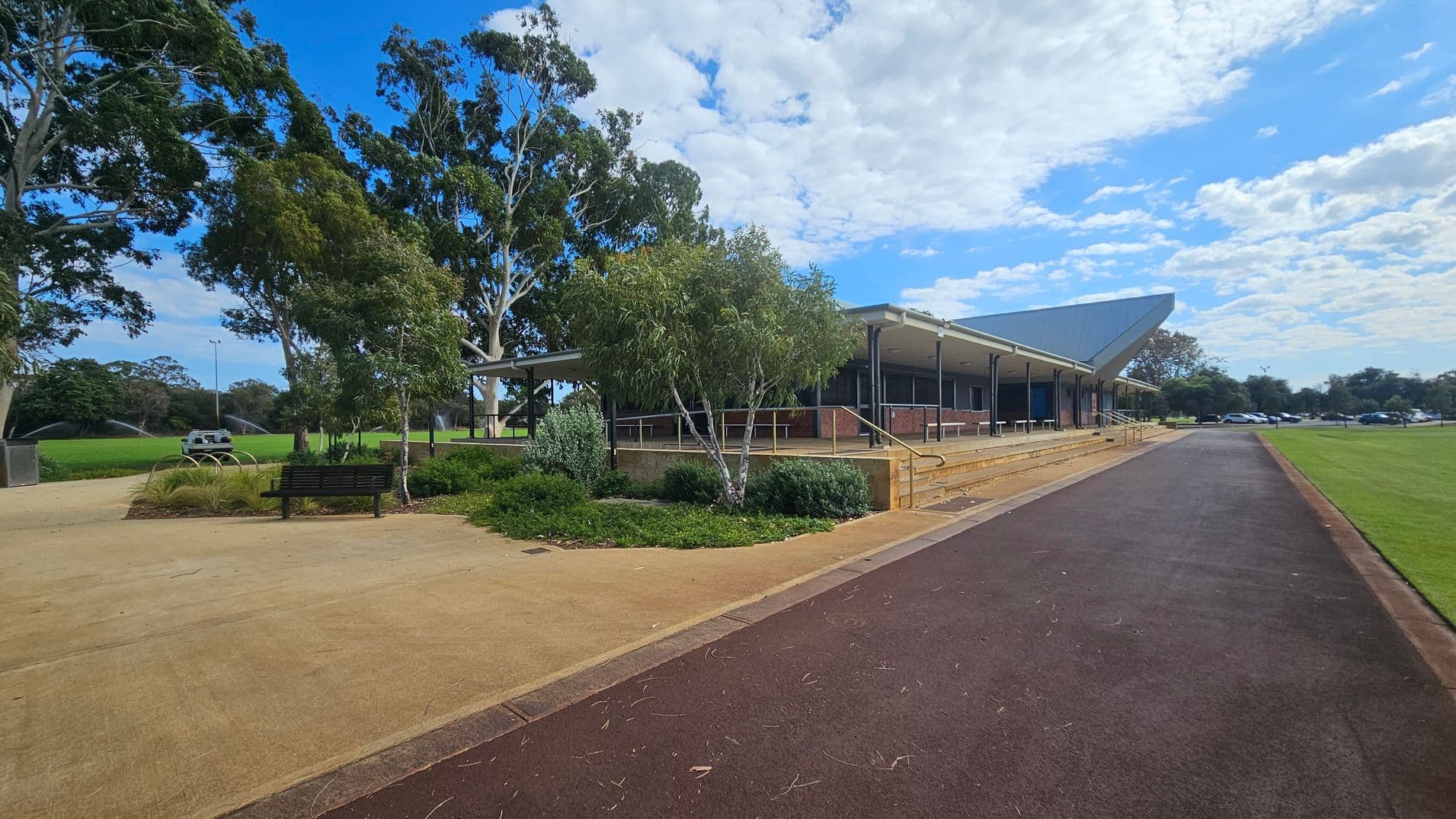 A paved path runs through the foreground, leading to a building on the right with a sloped roof and a series of steps. The building is surrounded by a landscaped area with shrubs and small trees. On the left, there is a bench and more greenery, with large trees in the background. A grassy field with sprinklers is visible to the left, and several parked cars can be seen in the distance on the right.