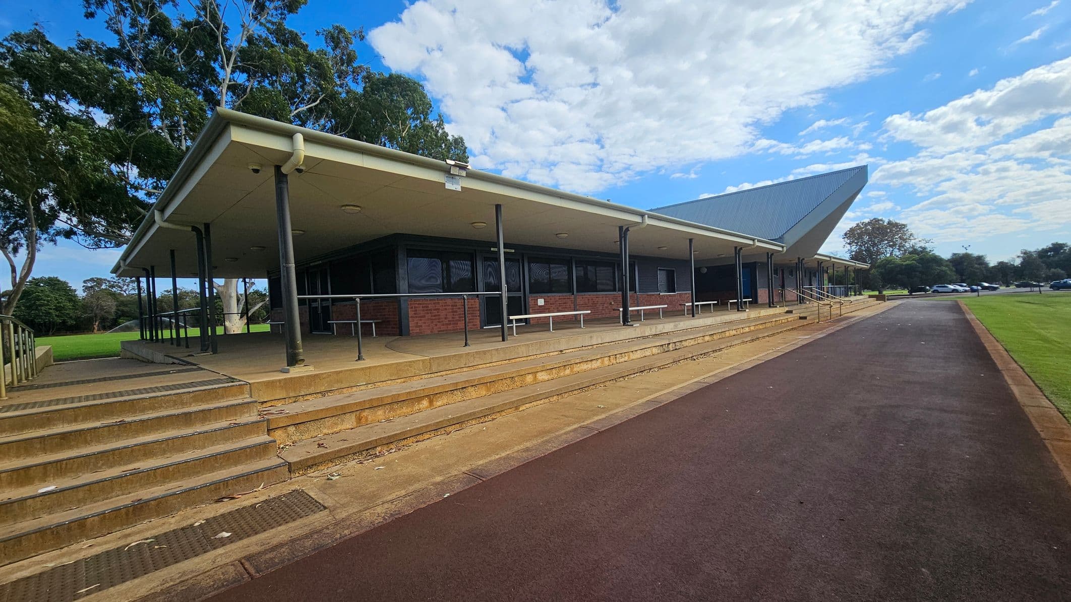 A modern building with a sloped roof and a covered walkway supported by black poles. The structure is made of brick with large windows. In the foreground, there are steps leading up to the entrance and a ramp with railings. The ground is paved with a reddish-brown path running alongside a grassy area. Trees are visible in the background, and several cars are parked in the distance. The sky is partly cloudy.
