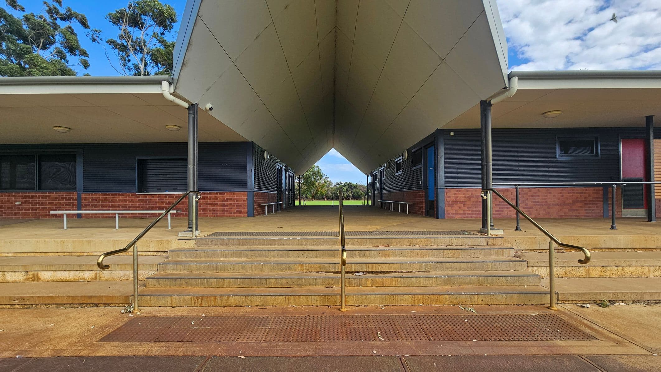 A symmetrical view of a building entrance with a wide, covered walkway leading to an open field in the background. The structure has a sloped roof supported by black columns. The walls are a combination of dark panels and red brick. Steps with metal railings lead up to the entrance. Benches are positioned along the walls on both sides. The ceiling is flat and light-coloured, with circular lights. Trees are visible in the distance beyond the walkway.