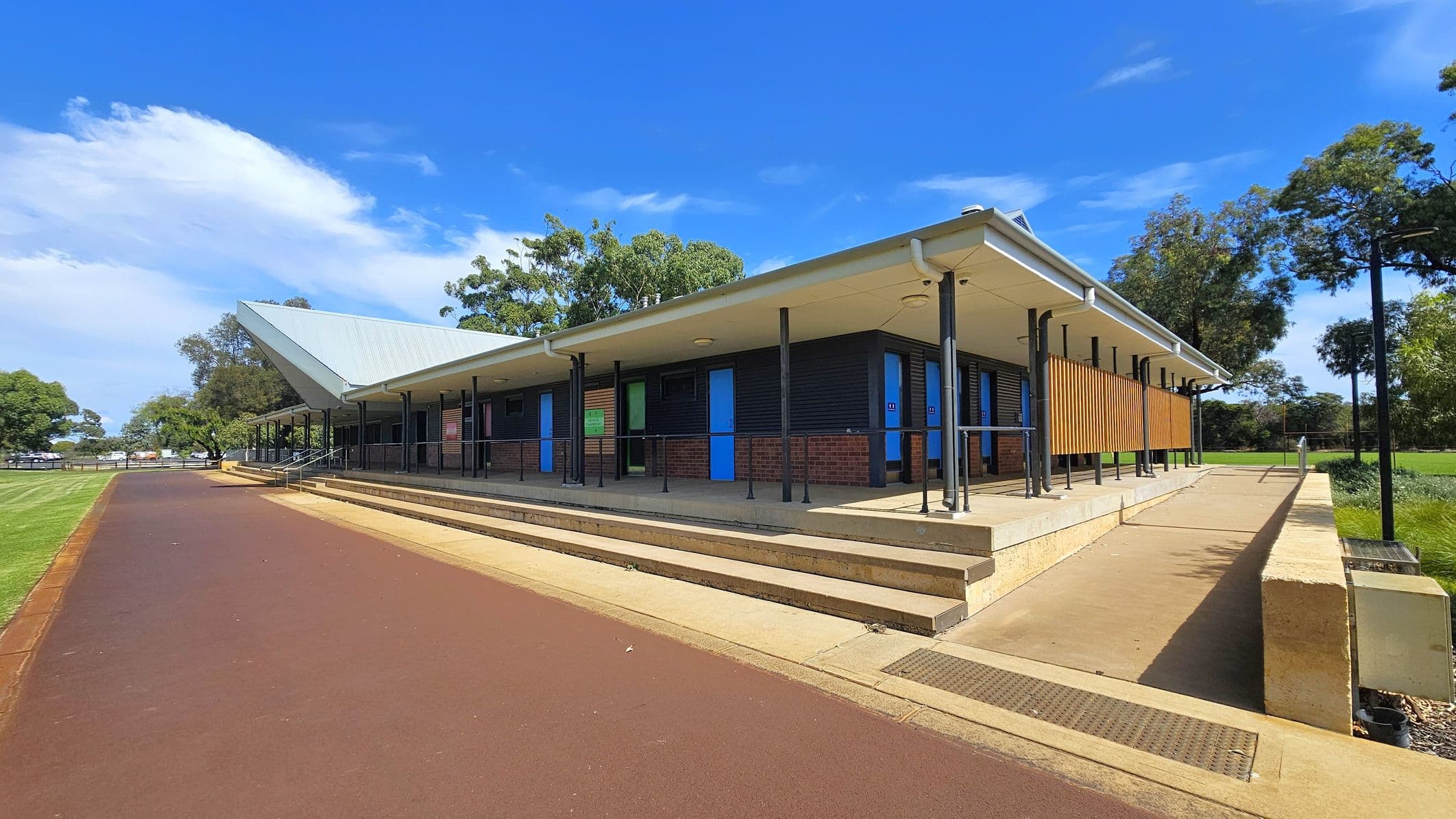 A modern building with a sloped metal roof and a covered walkway supported by black poles. The structure features a combination of brick and dark panelling, with bright blue doors. A series of steps and a ramp lead up to the entrance. On the left, a wide, reddish-brown path runs alongside a grassy area. Trees are visible in the background, providing a natural setting around the building.