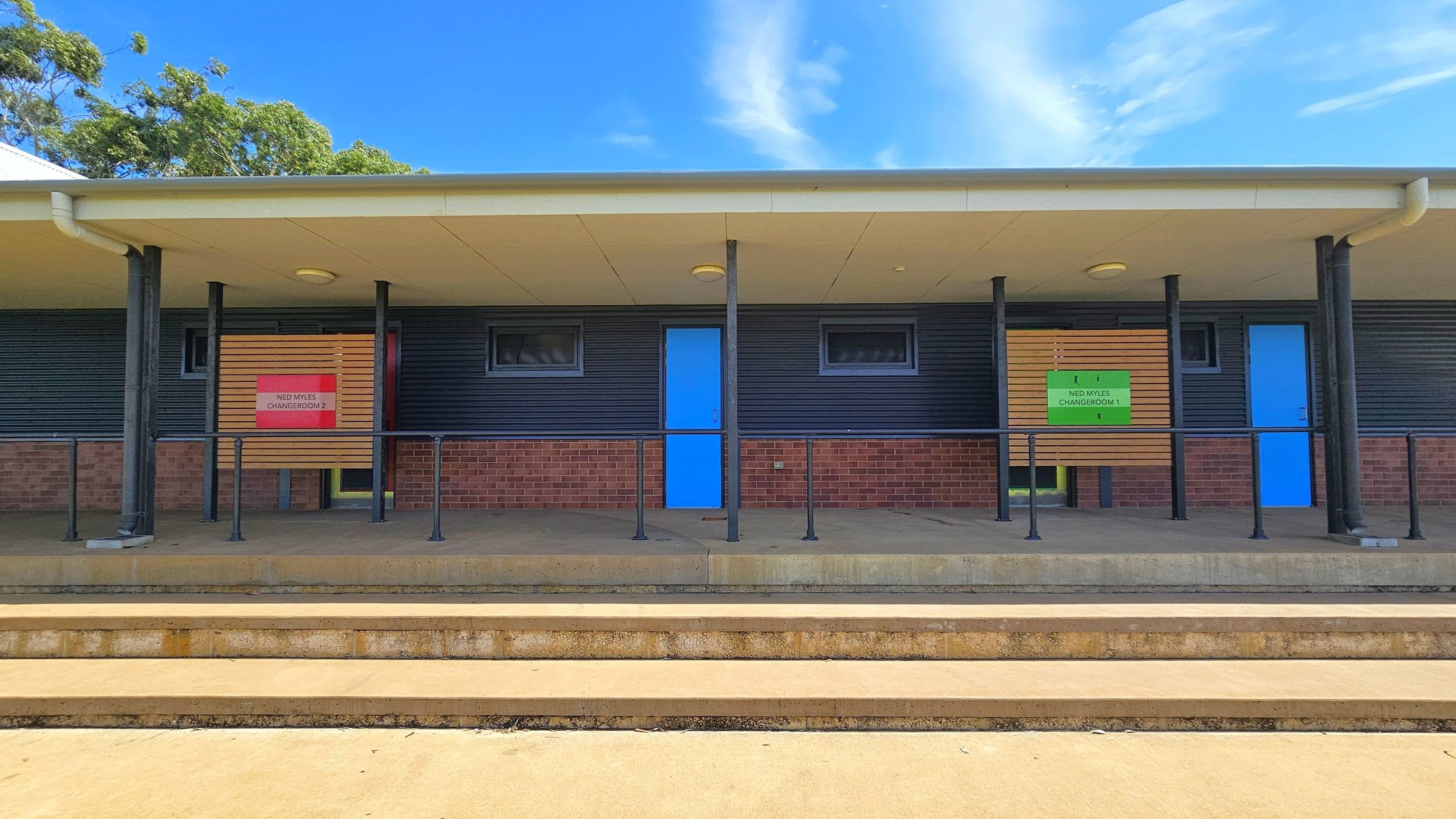 A building with a brick lower half and dark corrugated upper half. Two blue doors are centred between small windows. Signs above the doors read "Ned Myles Changeroom 2" on the left and "Ned Myles Changeroom 1" on the right. The roof is supported by black poles. The foreground features concrete steps leading up to the building. Trees are visible in the background.