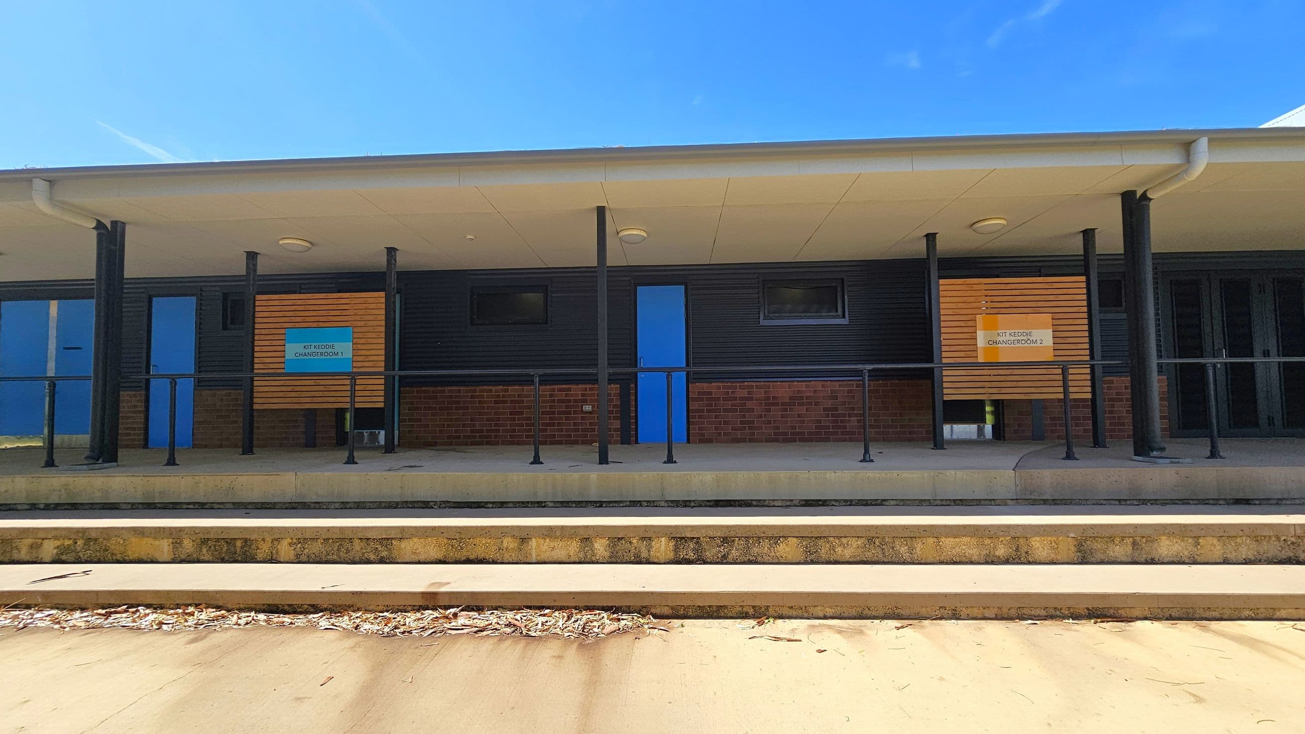 A building with a covered walkway supported by black poles. The facade features dark cladding and brickwork. Two blue doors are visible, each flanked by small rectangular windows. Signs above the doors read "Kit Keddie Changeroom 1" and "Kit Keddie Changeroom 2". The foreground has concrete steps leading up to the building, with some dried leaves scattered on the ground. The ceiling of the walkway is light-coloured with circular lights.
