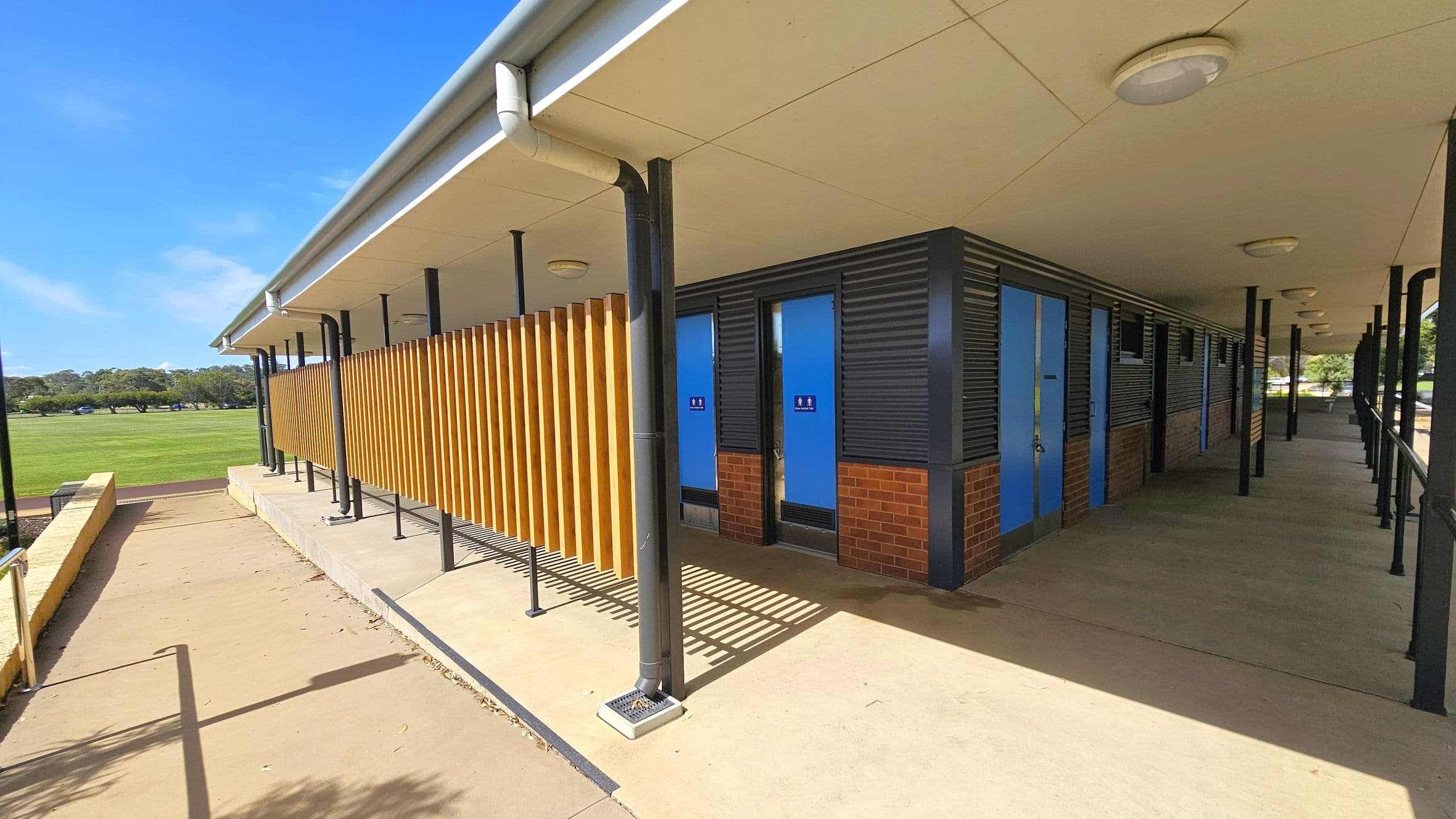 A building with a sloped roof and a series of blue doors on the right, likely public toilets, with signage indicating male and female facilities. The structure features a combination of brick and corrugated metal walls. Wooden slats run vertically along part of the exterior. The foreground has a concrete path with a metal railing, leading to a grassy field in the background. The ceiling has circular lights, and the area is supported by black metal poles.