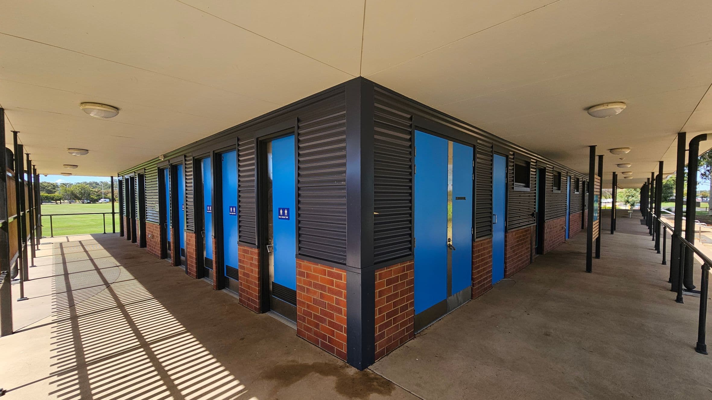 A building with a corner structure featuring blue doors and brick walls. The left side has a row of doors with signs, under a covered walkway with a concrete floor. The ceiling is white with round lights. The right side also has doors and windows, with black metal railings along the walkway. In the background, there is a grassy field and trees. Shadows from the railing create patterns on the floor.