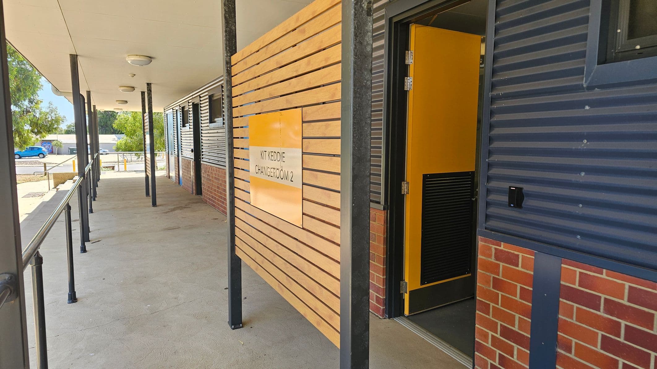 A corridor with a covered walkway featuring a series of black metal poles supporting the ceiling. On the right, a wooden panel with horizontal slats displays a sign for "Kit Keddie Changeroom 2" next to an open yellow door. The building has a combination of corrugated metal and brick walls. The floor is concrete with a greenish tint. In the background, there are trees and a blue car parked near some bollards.