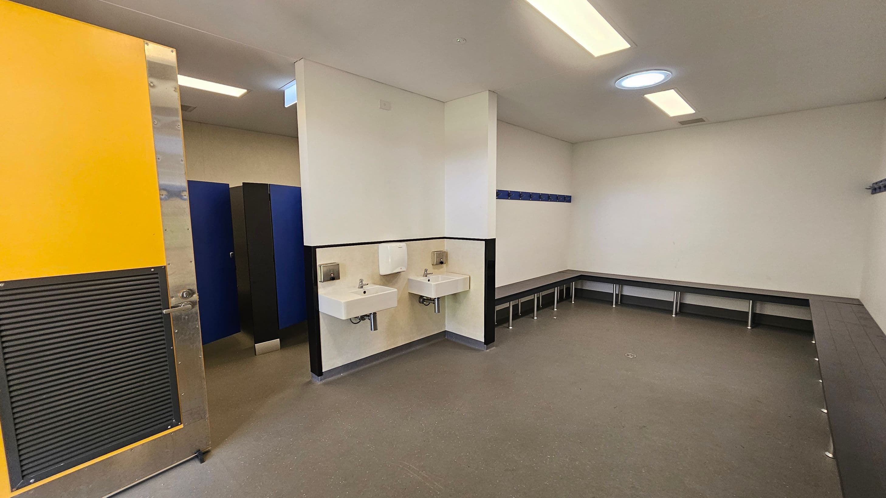 A changing room with a yellow door on the left, partially open, revealing blue lockers. In the centre, two white sinks are mounted on the wall with a paper towel dispenser above. On the right, a long bench runs along the walls, with hooks above for hanging items. The floor is grey, and the ceiling has rectangular lights and a circular skylight.