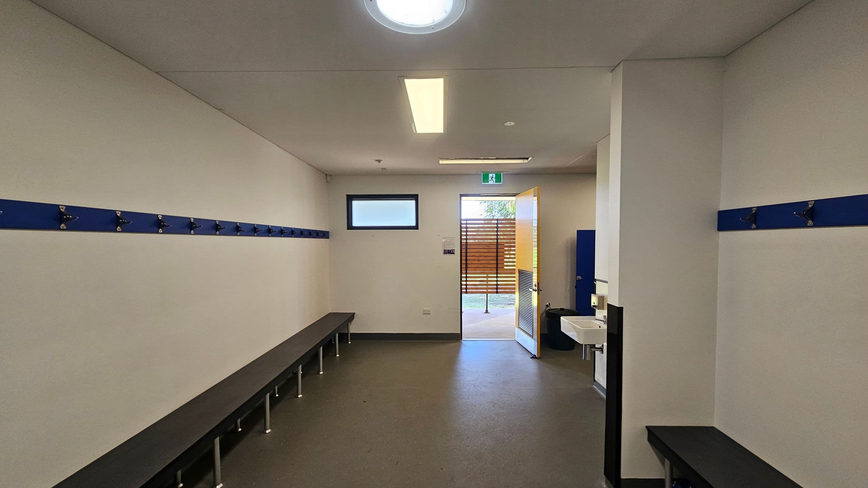 A changing room with white walls and a grey floor. On the left and right walls, there are blue hooks for hanging items, with long benches beneath them. In the middle, an open door leads outside, allowing natural light to enter. A small window is above the door. On the right, a white sink is mounted on the wall. The ceiling has a circular light fixture and a rectangular fluorescent light.