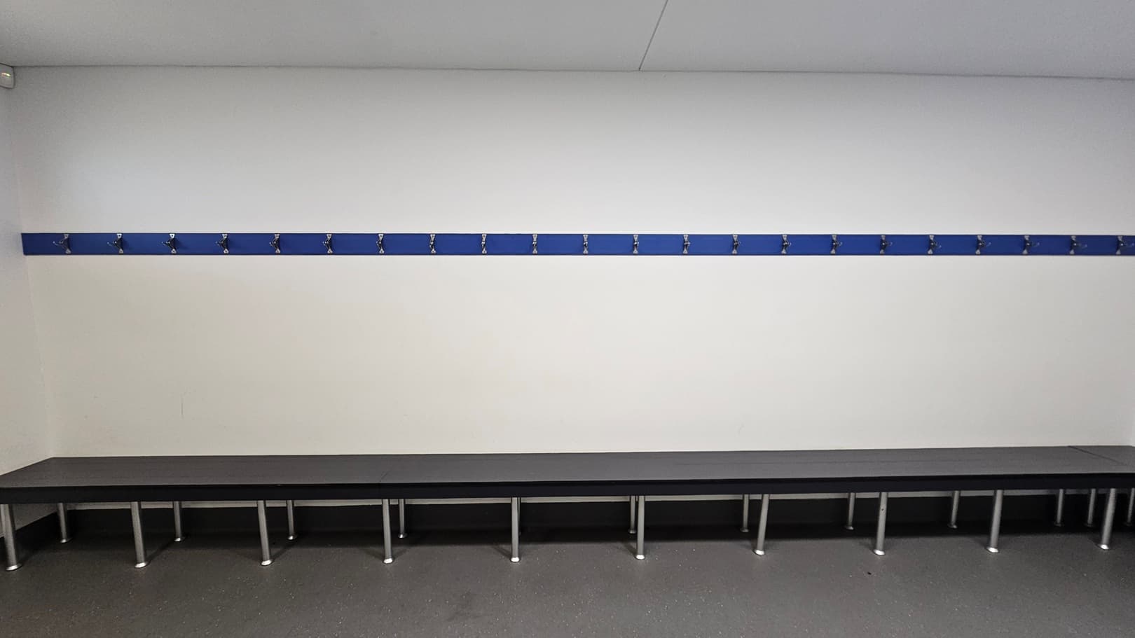 A room with a long, dark bench along the wall, supported by metal legs. Above the bench, a blue strip with multiple evenly spaced hooks is mounted on the white wall. The floor is grey and speckled, and the ceiling is plain white. The room appears to be a changing or locker room.