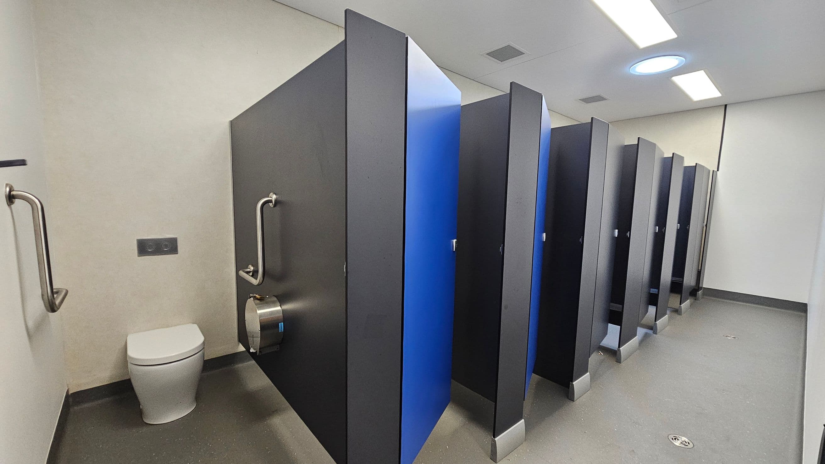A public restroom with a row of toilet cubicles on the right, featuring dark grey partitions and blue doors. On the left, there is a single toilet with grab bars on either side and a toilet paper dispenser attached to the partition. The floor is grey with a drain visible, and the ceiling is white with rectangular and circular lights.