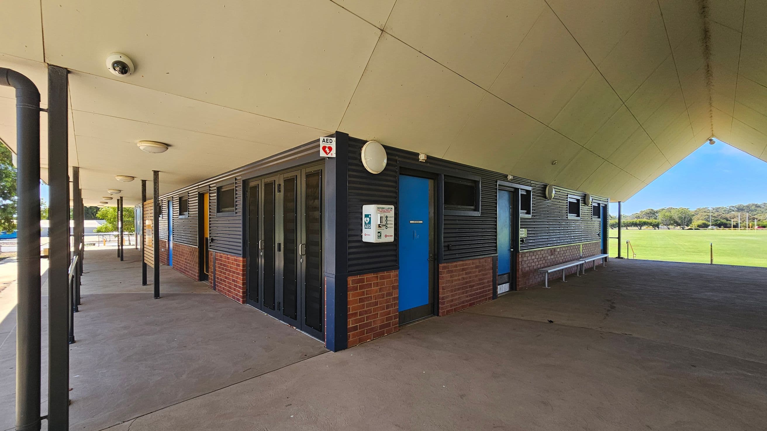 A building with a sloped roof and a covered walkway. The exterior walls are a combination of dark panels and red brick. A blue door is visible on the right, with a sign for an AED (Automated External Defibrillator) mounted nearby. Several windows are along the wall. The ceiling is light-coloured with circular lights. The floor is concrete, and there are metal benches along the wall. In the background, there is a grassy field with trees.