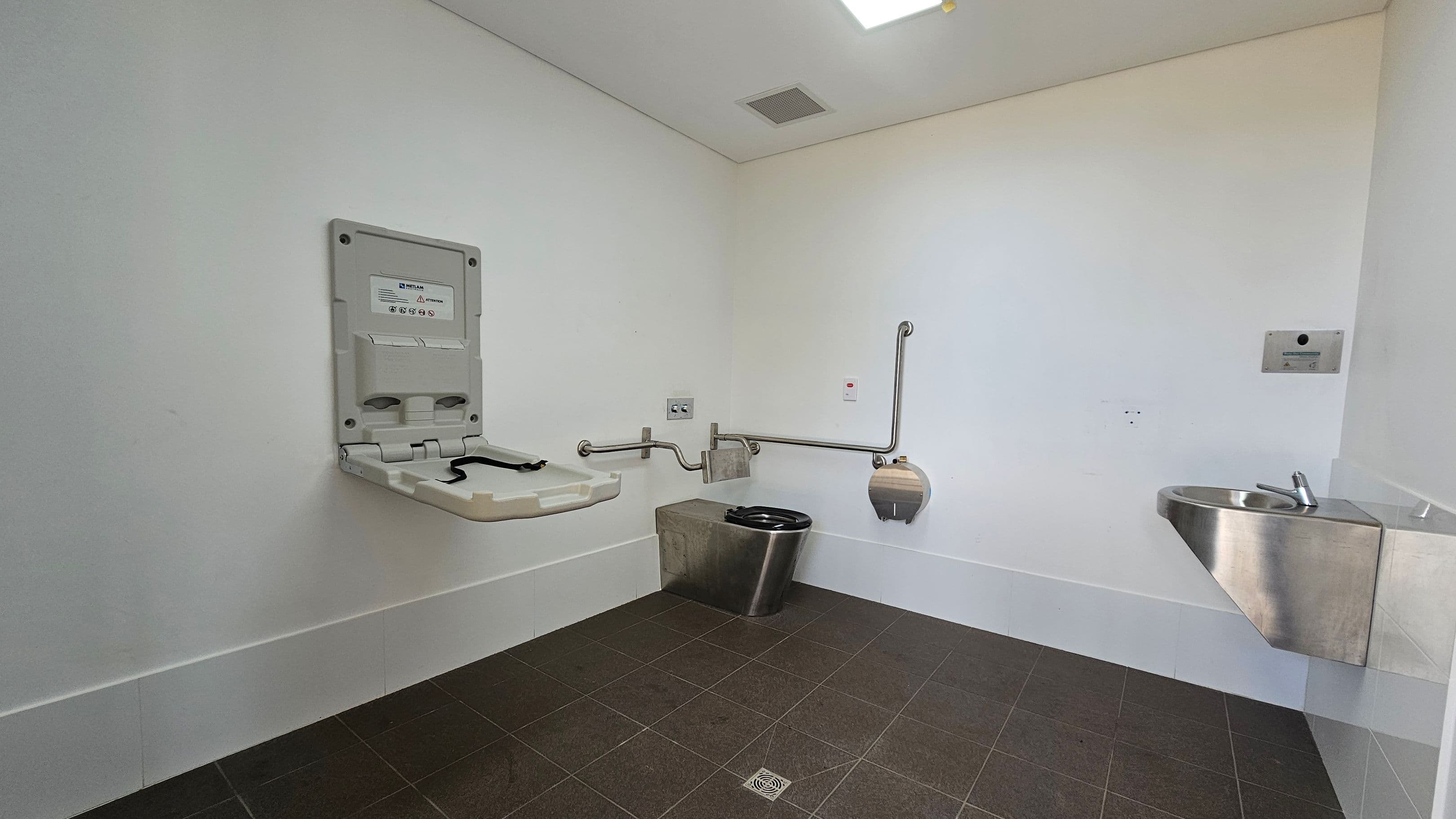 A public restroom with a baby changing station on the left wall. In the centre, there is a stainless steel toilet with grab bars on both sides and a toilet paper dispenser. On the right, a small stainless steel sink is mounted on the wall. The floor is tiled in dark grey, and the ceiling is plain white with a vent and a light fixture.