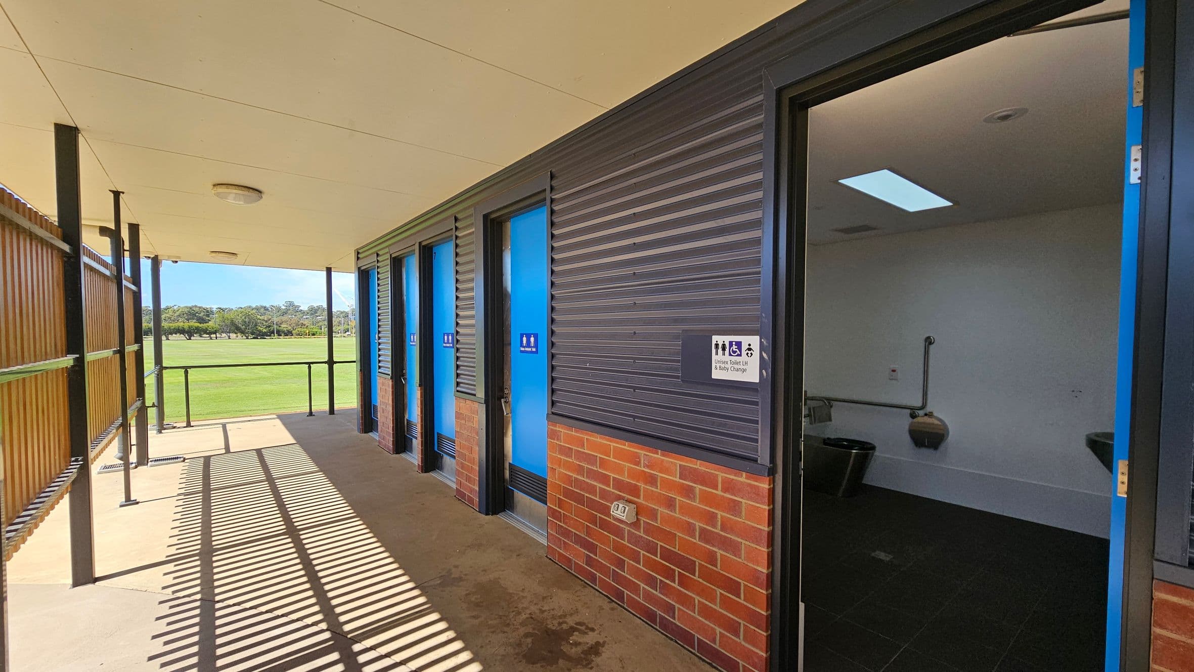 A row of public toilets with blue doors is on the right, set against a wall with dark corrugated panels and a brick base. One door is open, revealing a white interior with a toilet and grab bars. A sign indicates unisex, disabled, and baby change facilities. On the left, a wooden slatted fence casts shadows on the concrete floor. The ceiling is light-coloured with circular lights. In the background, a grassy field and trees are visible.