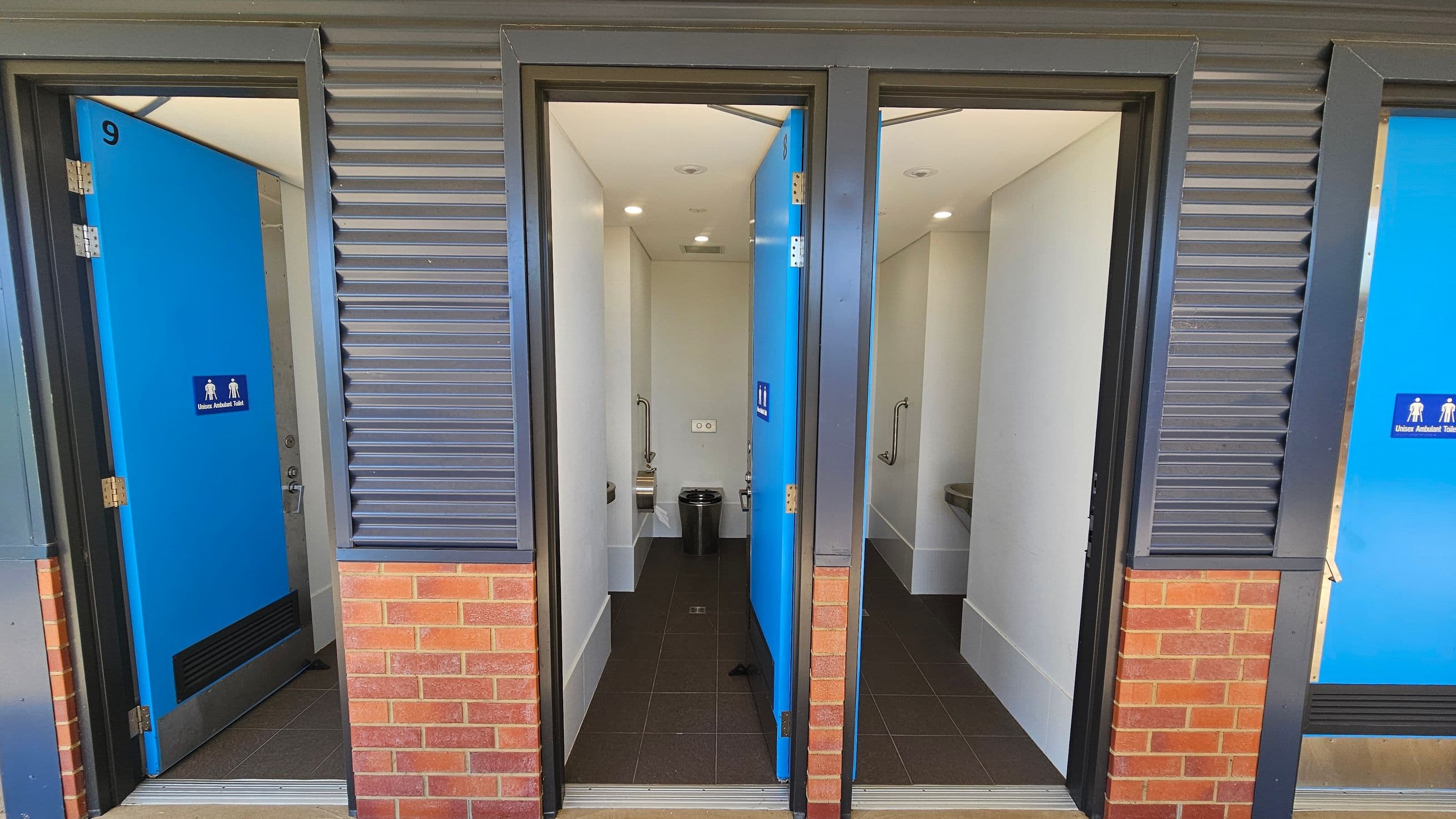 Three public toilet cubicles with blue doors, each labelled as unisex ambulant toilets. The doors are partially open, revealing white interiors with grab bars and waste bins. The exterior wall features a combination of grey metal cladding and red brick. The floor inside the cubicles is dark tiled, and the ceiling is white with recessed lighting.