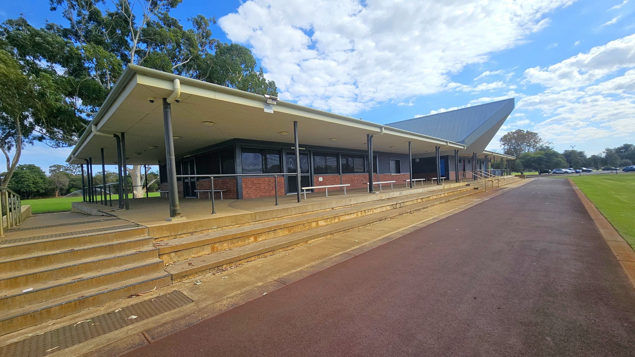 A building with a sloped roof and a covered walkway supported by black poles. The structure is made of brick with large windows. Steps lead up to the entrance, and there are benches along the walkway. On the left, there are trees with green foliage. On the right, a paved path runs alongside a grassy area, with parked cars visible in the distance. The sky is partly cloudy.