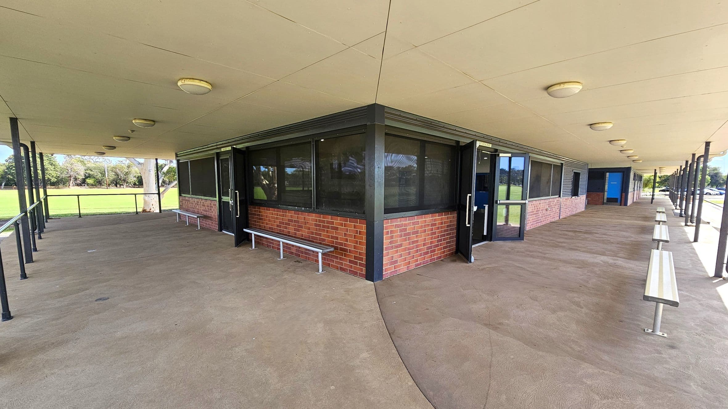 A covered outdoor area with a beige ceiling featuring round lights. The floor is concrete, and there are metal benches along the walls. The building has brick walls with large windows and glass doors. On the left, there's a grassy field with trees in the background. On the right, the structure extends with more benches and a blue door visible. The area is supported by black metal poles.