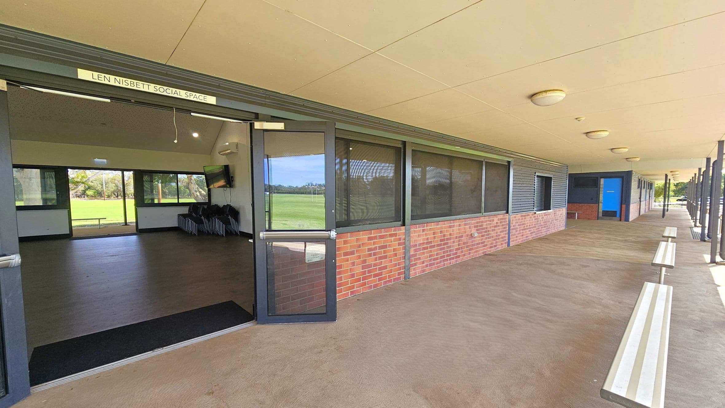 A covered outdoor walkway with a beige ceiling and concrete floor. On the left, a door is open to a room labelled "Len Nisbett Social Space," revealing a dark floor and windows overlooking a grassy field. The right side features a brick and metal-panelled wall with several windows and a blue door. Benches are positioned along the walkway, and round ceiling lights are evenly spaced above.