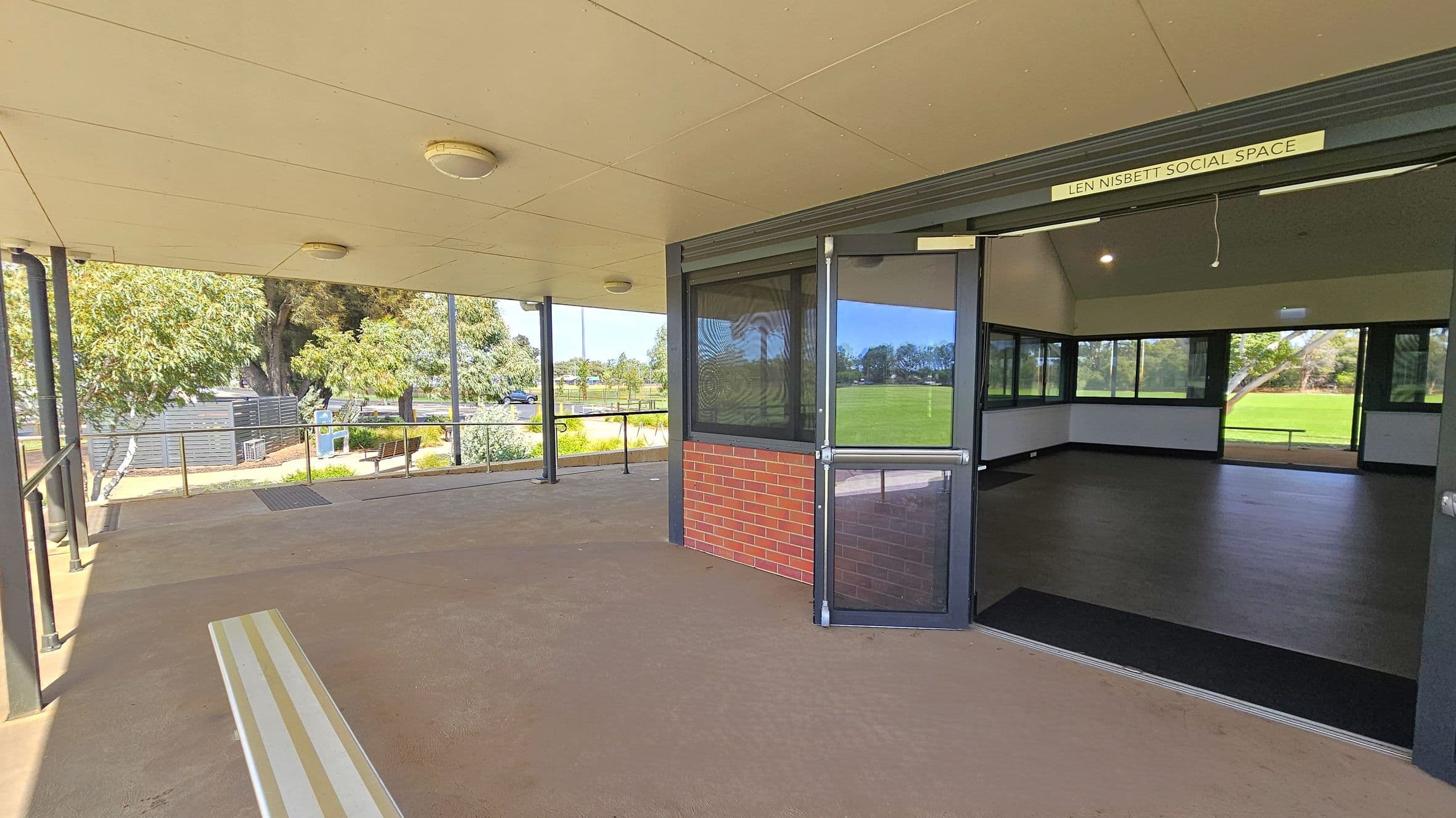 A covered outdoor area with a beige ceiling and several round lights. On the left, there are trees and a pathway with benches. The entrance to the "Len Nisbett Social Space" is on the right, featuring a glass door and brick wall. Inside, the room has dark flooring and large windows overlooking a grassy field. The floor outside is a light brown concrete.