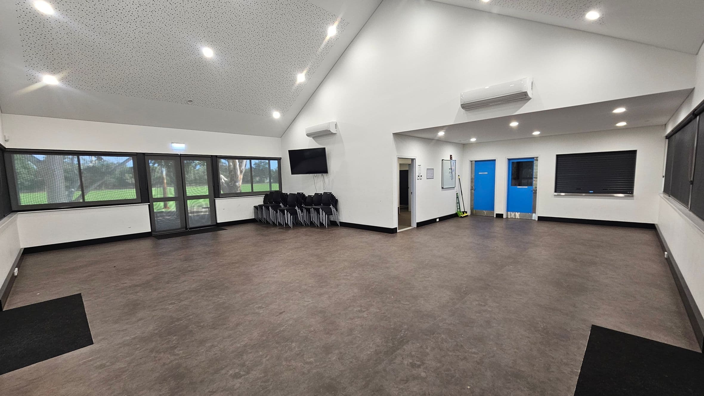 A spacious room with a brown floor and a white, perforated ceiling with recessed lights. On the left, large windows and a glass door offer a view of greenery outside. In the centre, a stack of black chairs is placed against the wall beneath a mounted TV. On the right, two bright blue doors are visible, along with a closed shutter window. A whiteboard and cleaning tools are mounted on the wall near the doors. The room has a clean and organised appearance.