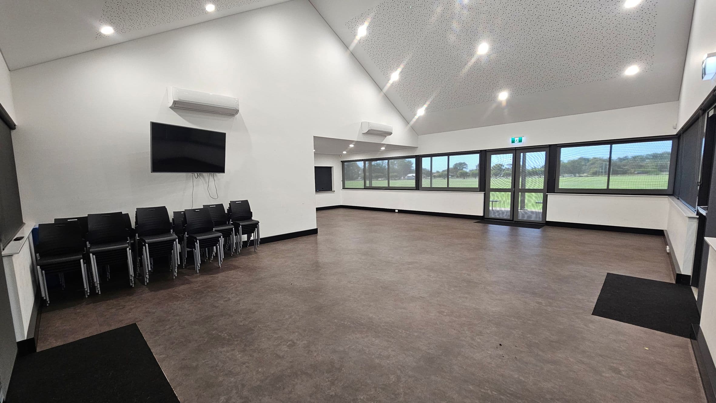 A spacious room with a high, sloped ceiling featuring recessed lights. On the left, there is a stack of black chairs beneath a wall-mounted television and an air conditioning unit. The floor is a mix of brown and purple tones. The right side has large windows and a glass door, offering a view of a grassy field outside. The walls are white, and there is another air conditioning unit above the windows.