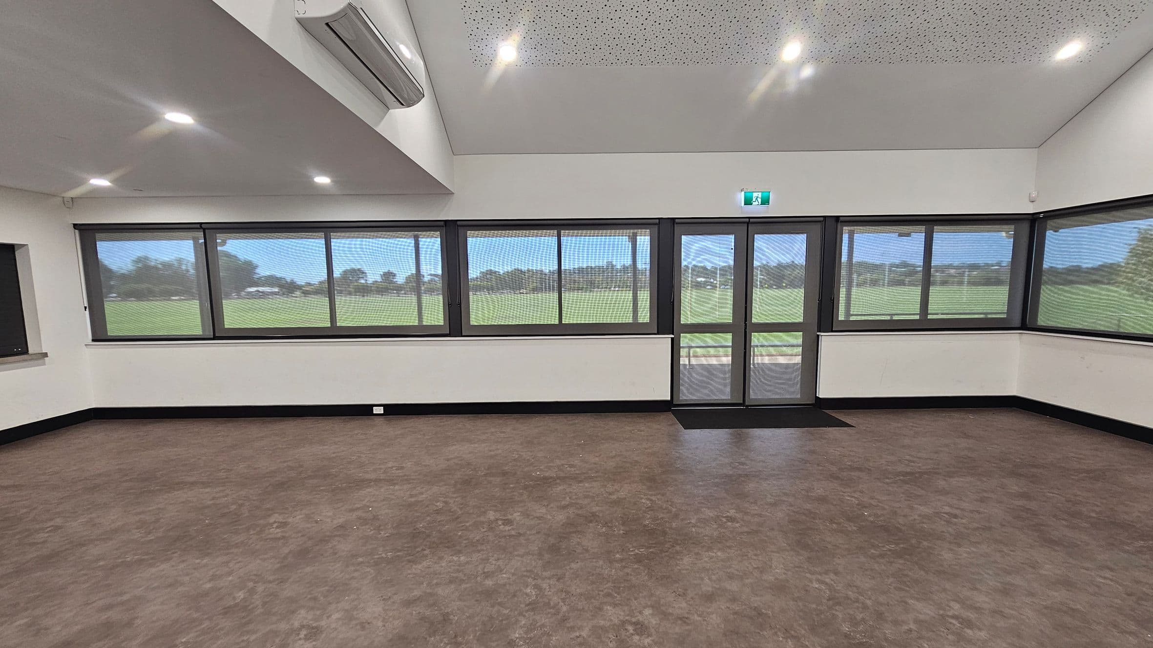A room with large windows and a glass door on the right, offering a view of a grassy field outside. The walls are white, and the floor is brown with a smooth finish. The ceiling is white with recessed lighting and an air conditioning unit on the left. The windows have a mesh screen, and there is a black skirting board along the bottom of the walls. An emergency exit sign is above the door.
