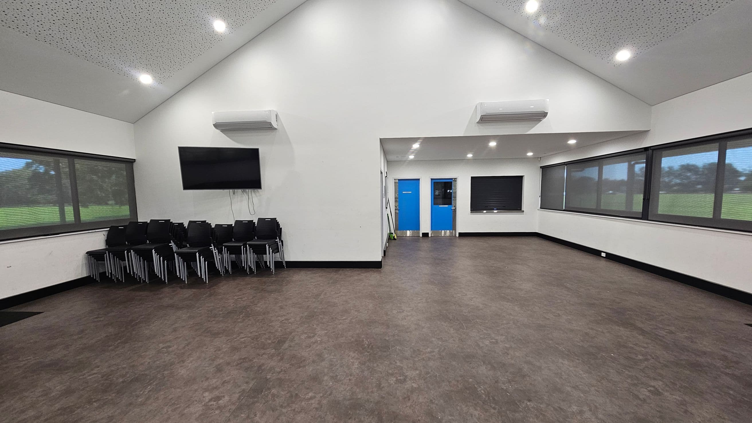A spacious room with a high, sloped ceiling featuring recessed lights. On the left, there is a stack of black chairs beneath a wall-mounted television. The right side has large windows with blinds, offering a view of a grassy area outside. In the centre, two blue doors are visible, with a small window between them. The floor is a mix of brown and purple tones, and there are air conditioning units mounted on the walls.