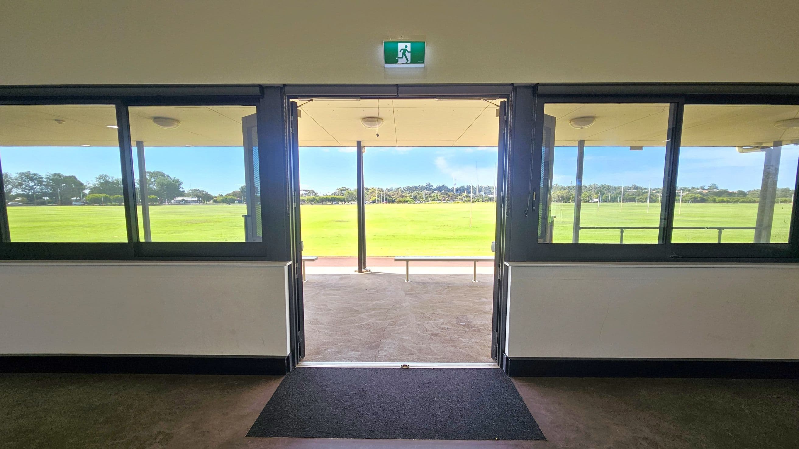 A view from inside a building looking out through a glass door. On either side of the door are large windows. Above the door is an emergency exit sign. Outside, there is a covered area with a bench and a wide expanse of green grass, possibly a sports field. Trees and a few structures are visible in the distance. The floor inside is carpeted near the entrance, transitioning to a hard surface outside. The ceiling inside is plain with a light fixture.