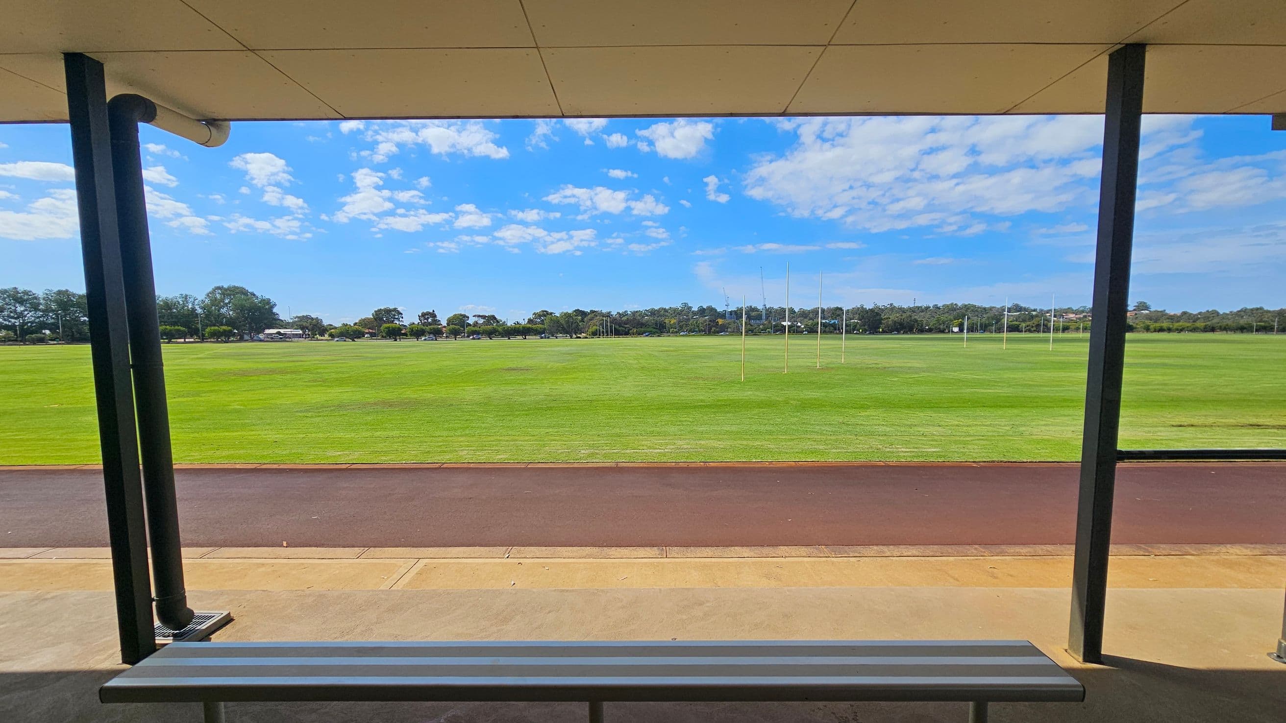 A sports field with green grass and several goalposts in the distance. In the foreground, a sheltered area with a metal bench is visible, supported by dark vertical poles. The ground is a mix of concrete and a reddish-brown track. Trees and a few buildings are visible on the horizon. The ceiling of the shelter is light-coloured and flat.
