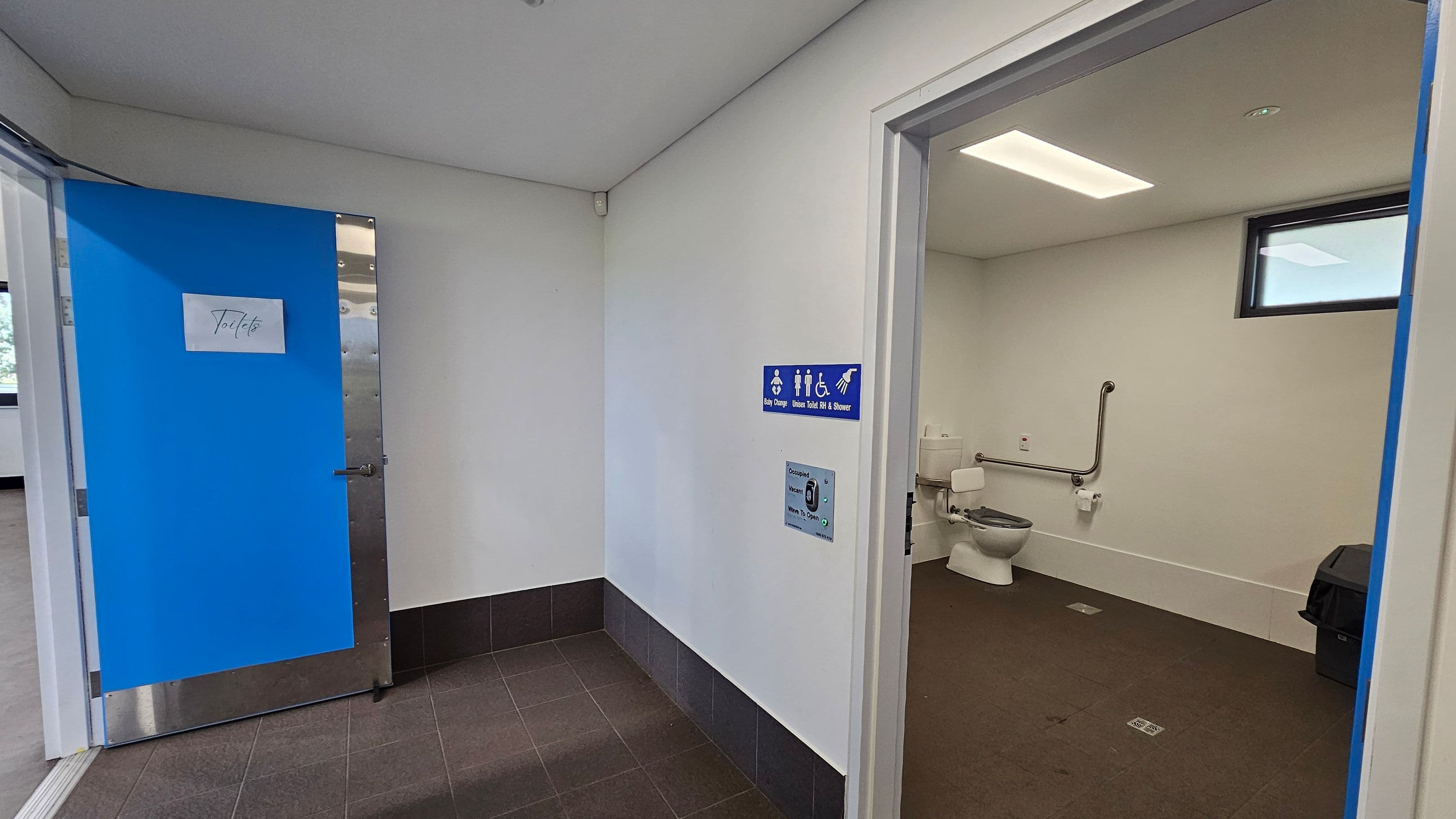 A public toilet area with a blue door on the left, labelled "Toilets". The floor is tiled in dark grey. On the right, an open door reveals an accessible bathroom with a toilet, grab bars, and a waste bin. A blue sign indicates facilities for baby change, toilets, and a shower. The ceiling is white with a rectangular light fixture, and a small window is visible in the bathroom.