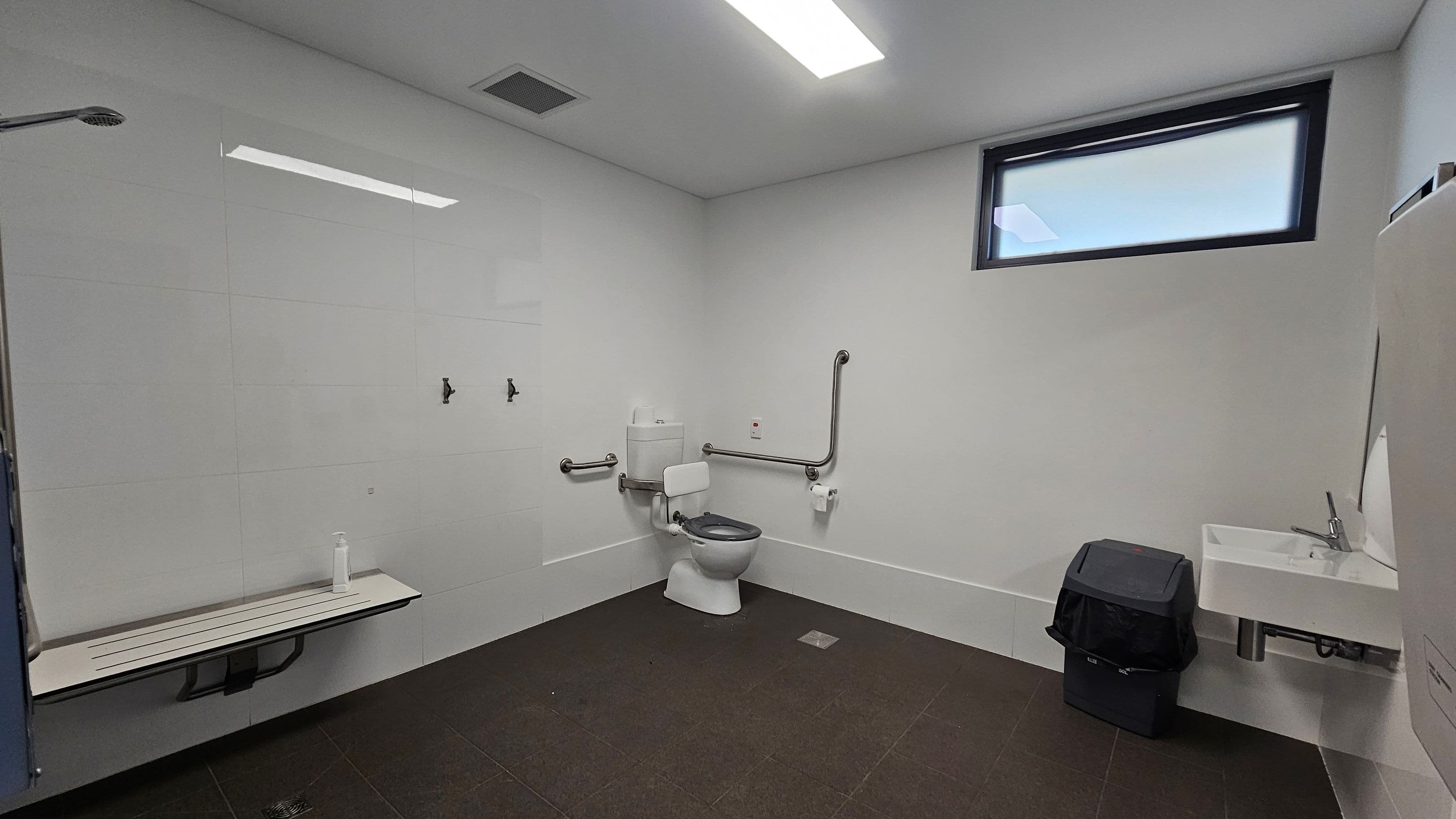 A spacious, accessible bathroom with white tiled walls and a dark tiled floor. On the left, a foldable bench with a bottle on it. In the centre, a toilet with support rails on both sides and a toilet paper holder. Above the toilet, a small window provides natural light. On the right, a white sink with a tap, and a black bin underneath. The ceiling has a rectangular light fixture and a vent.