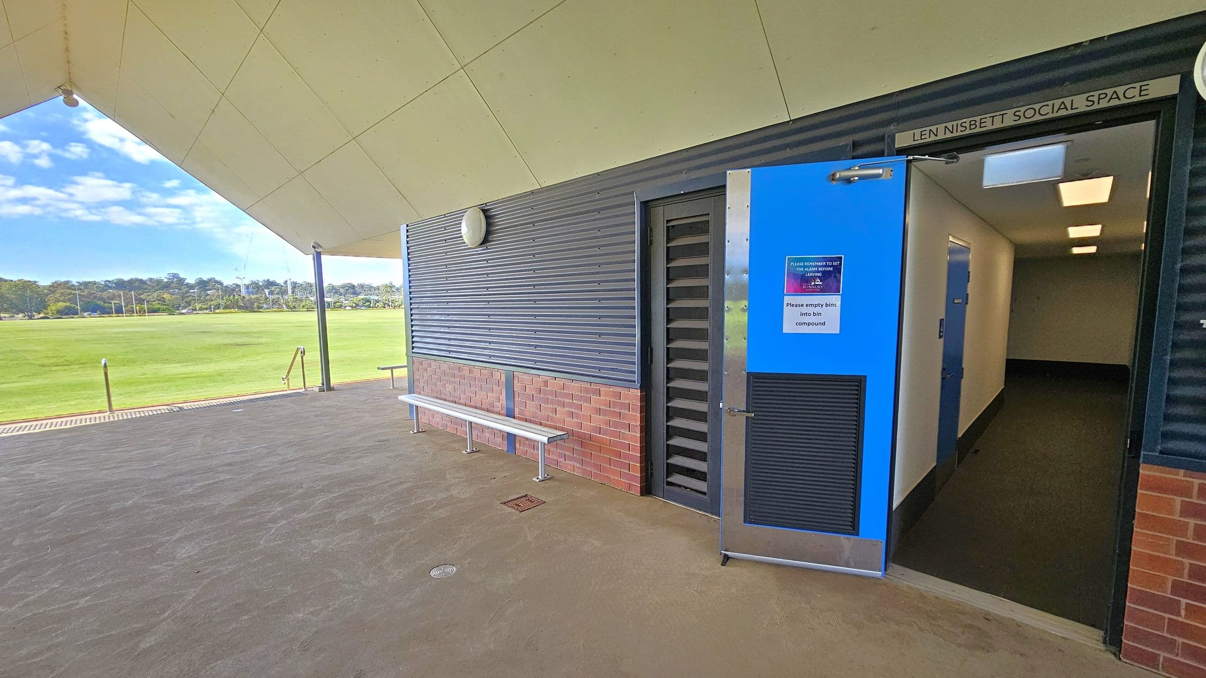 A covered outdoor area with a view of a grassy field in the background. On the right, a blue door is open, leading into a hallway with white walls and a dark floor. Above the door, a sign reads "Len Nisbett Social Space." The exterior wall is a combination of corrugated metal and brick, with a white bench attached. The ceiling is angular and light-coloured, supported by a black pole. The floor is concrete, and there are metal railings leading down to the field.