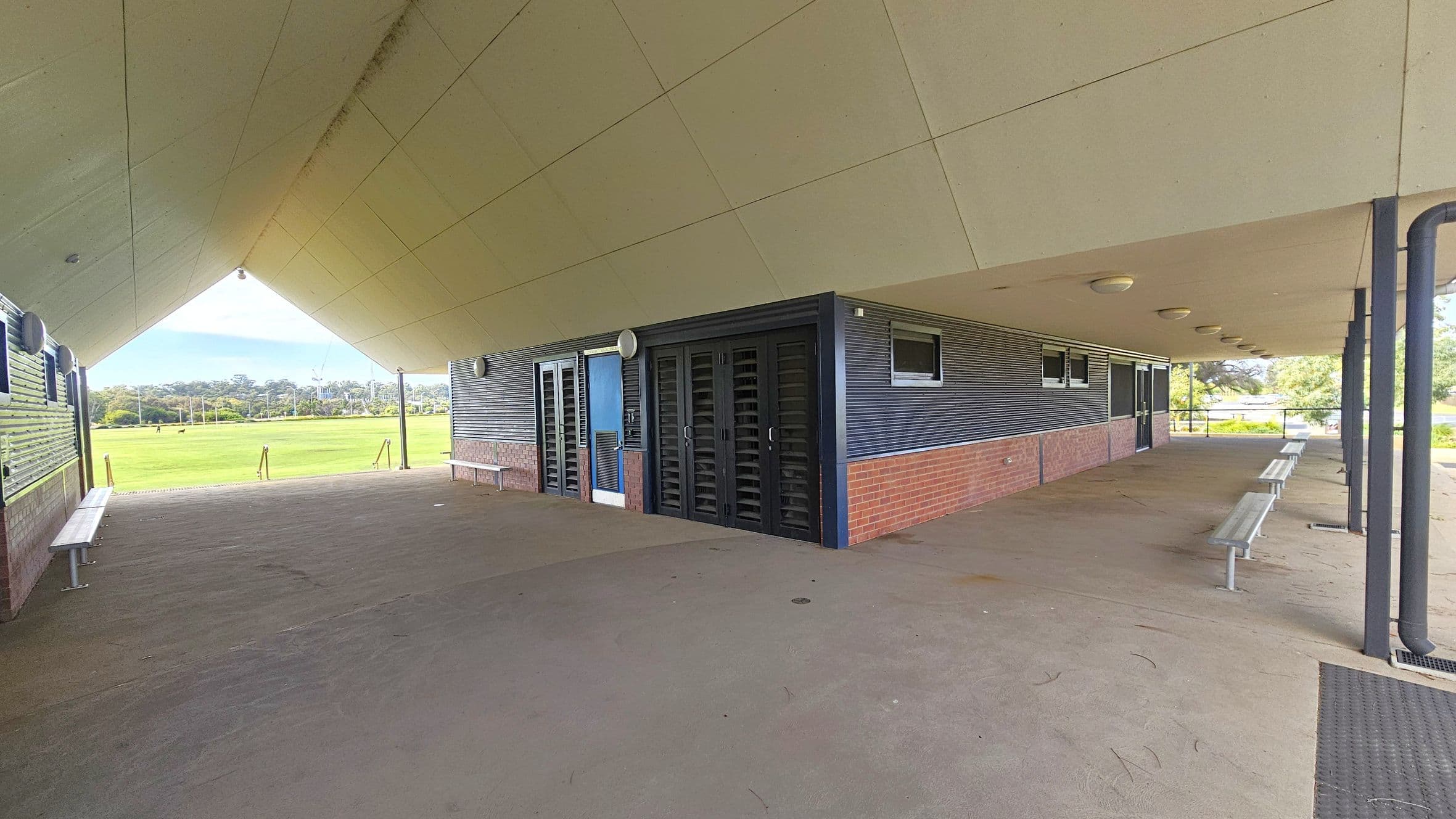 A covered outdoor area with a sloped ceiling and a concrete floor. On the right, there are metal benches and a structure with brick and corrugated metal walls, featuring doors and small windows. The left side opens to a grassy field with trees in the distance. The ceiling has several round lights, and the area is supported by metal columns.