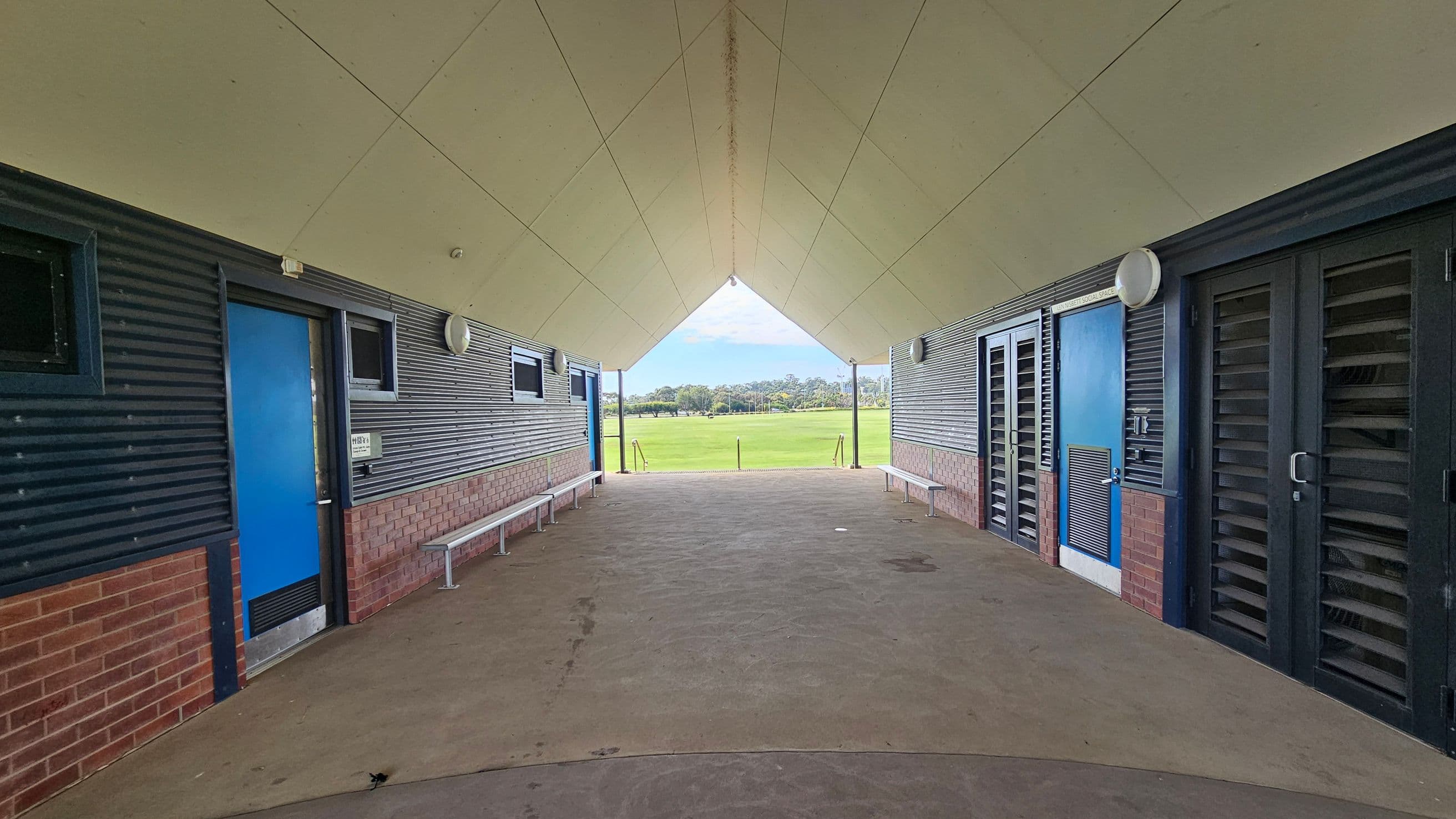 A covered walkway with a triangular ceiling leads to an open grassy field in the background. On the left, there is a blue door and a bench along a wall with corrugated metal and brick. On the right, similar walls feature a blue door and louvred panels. The floor is concrete, and the ceiling is light-coloured with a central seam.