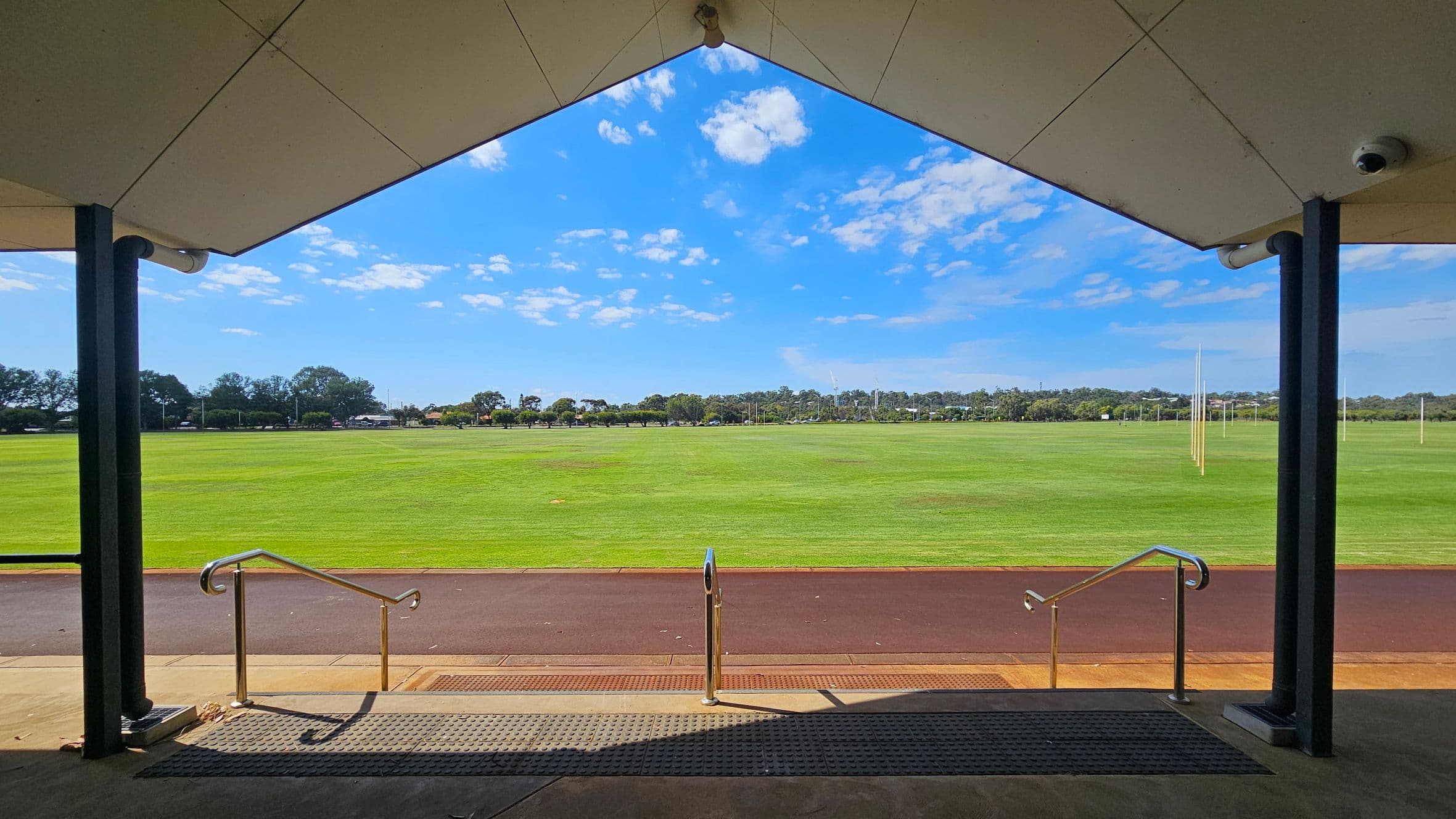 A view from under a sheltered area with a triangular roof, supported by black columns. In the foreground, metal railings lead down steps to a paved path. Beyond the path lies a large, well-maintained grassy field with several tall goalposts. The background features a line of trees and some distant structures. The ceiling of the shelter is light-coloured, and the floor is concrete with a textured mat near the steps.
