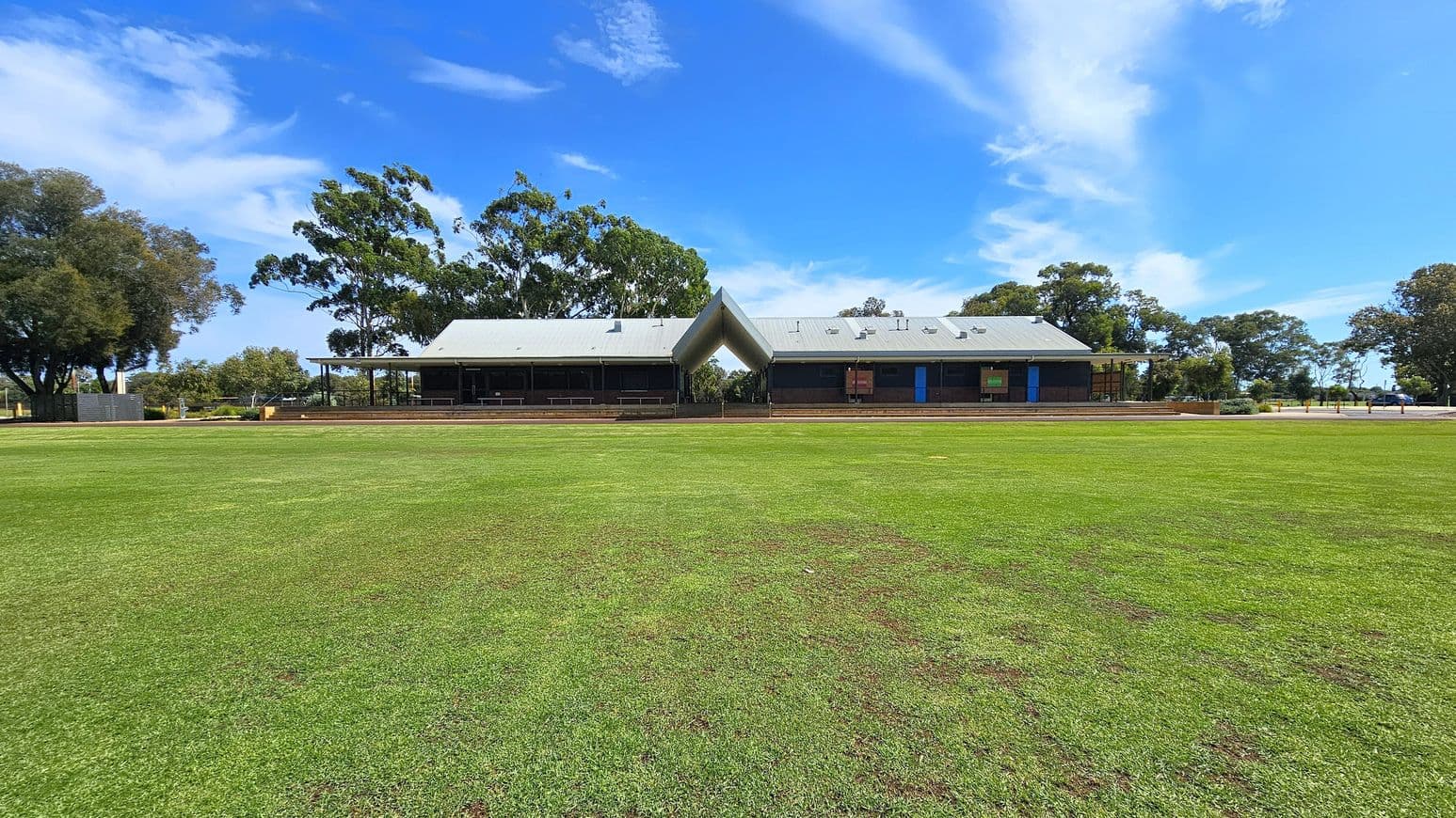 A large, open grassy field in the foreground leads to a modern building in the centre. The building has a distinctive V-shaped roof and is flanked by trees on both sides. The structure features a covered veranda and large windows. The sky is clear with some clouds, and the area is surrounded by more trees, creating a natural backdrop.