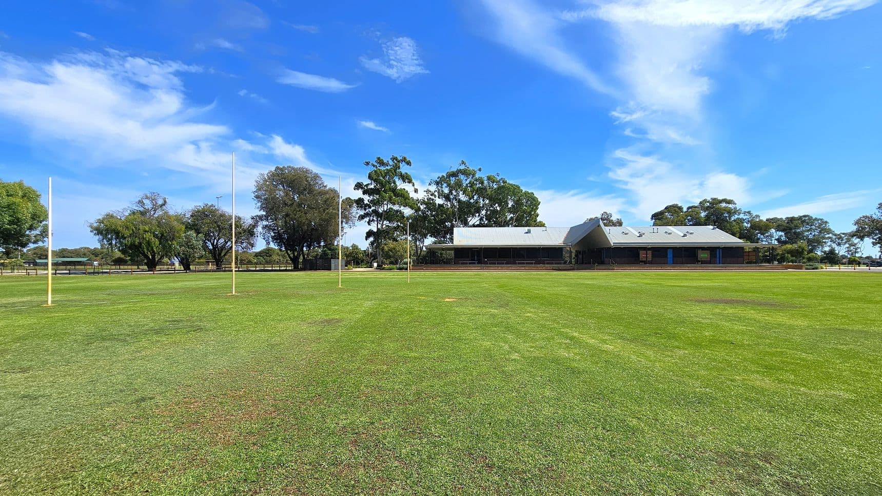 A grassy sports field with four tall goalposts in the centre. In the background, there is a building with a grey roof and a veranda, surrounded by several trees. The field is expansive, with a well-maintained green surface. The sky is clear with some clouds.