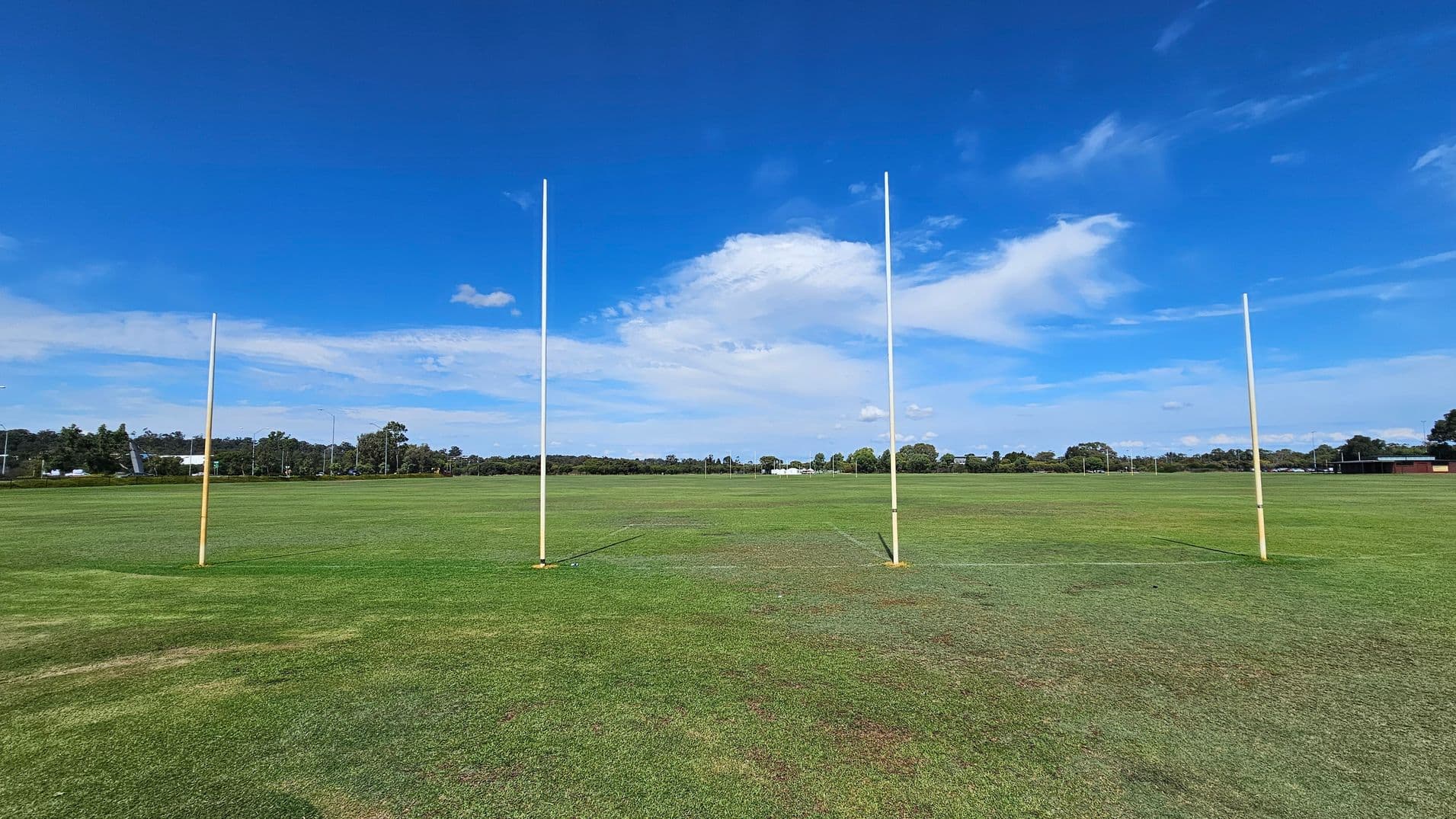 Four tall goalposts stand on a large, grassy field. The grass is a mix of green and brown patches. In the background, there are trees and some low buildings. The sky is clear with a few clouds.