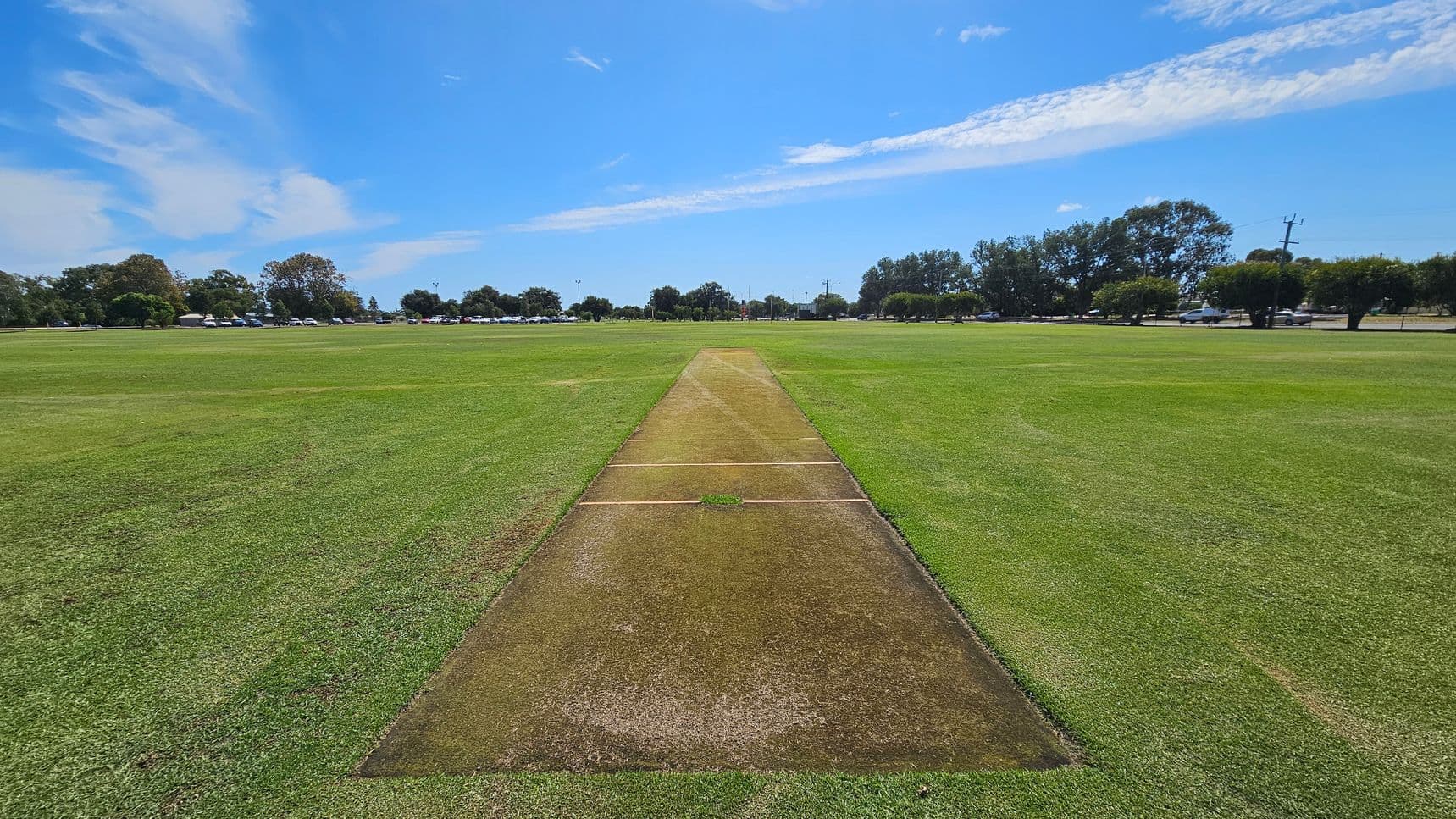 A cricket pitch is in the centre of a large grassy field. The pitch is a rectangular strip with a worn, brown surface. Surrounding the pitch is a well-maintained green outfield. In the background, there are trees and a line of parked cars along a road. The sky is blue with some clouds.