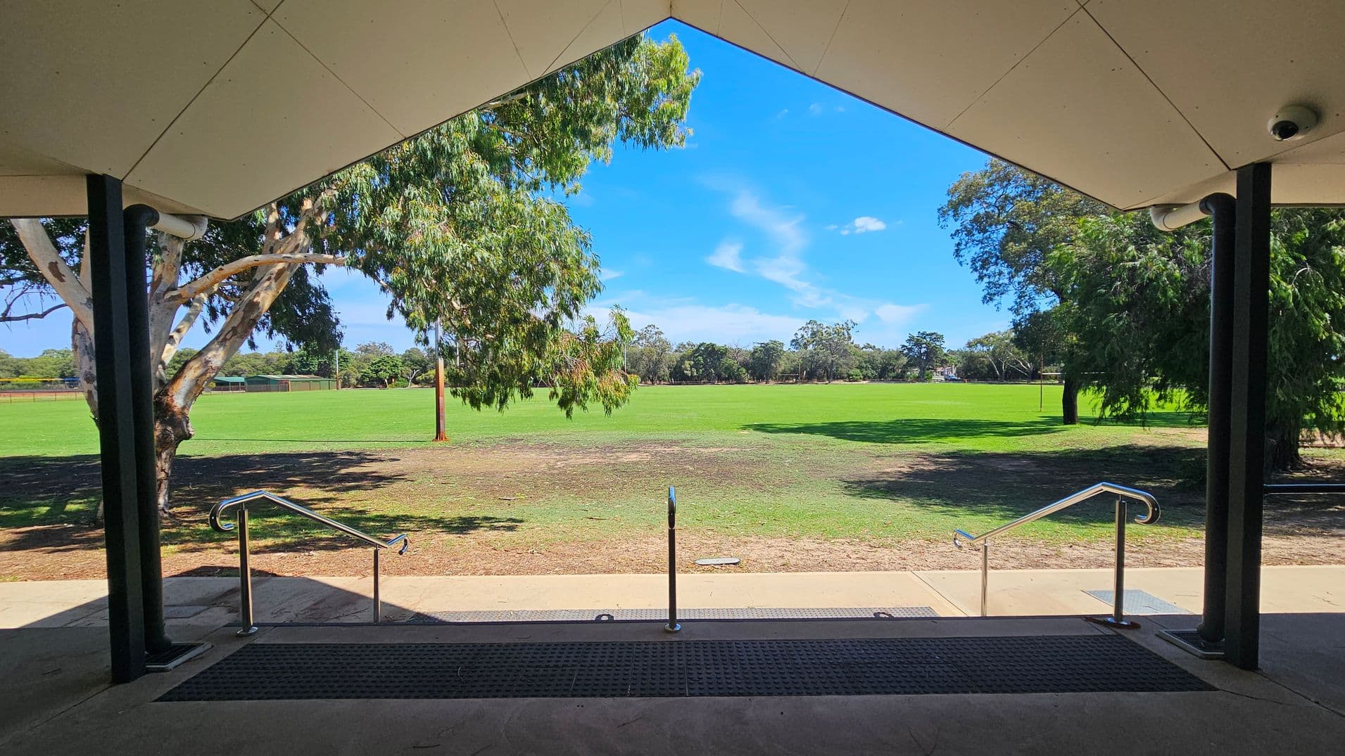 A view from under a sheltered area with a triangular ceiling, supported by black columns. In the foreground, there are steps with metal railings leading down to a grassy field. The field is expansive and bordered by trees on both sides. A large tree is visible on the left, and another on the right, with more trees in the distance. The ground near the steps is a mix of grass and bare patches.