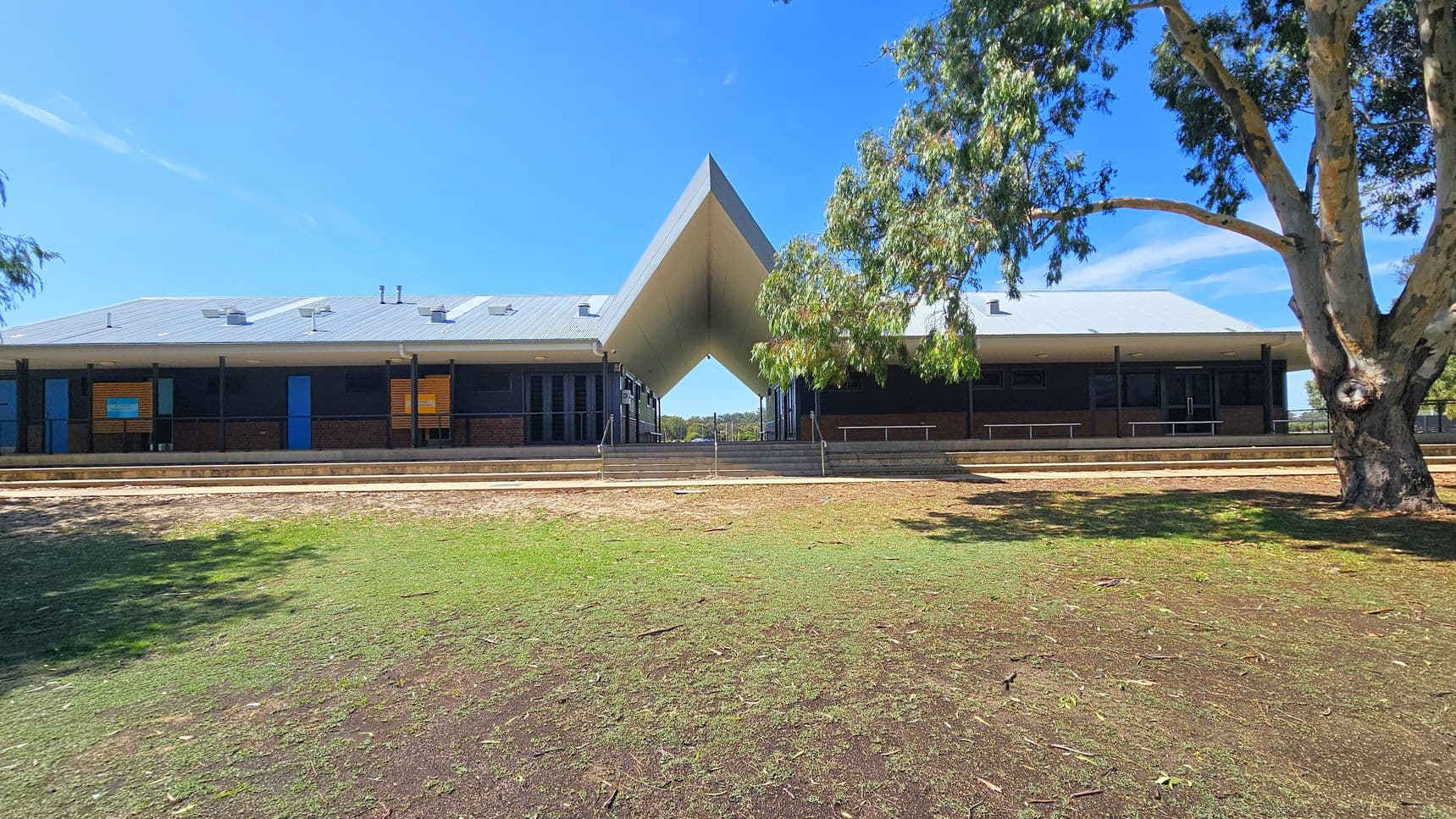 A building with a distinctive V-shaped roof structure in the centre. The building has a grey roof and dark walls with several blue doors and windows. In the foreground, there is a grassy area with patches of dirt and a large tree on the right. The background shows a clear blue sky. The building is elevated with steps leading up to it, and there are railings along the front.