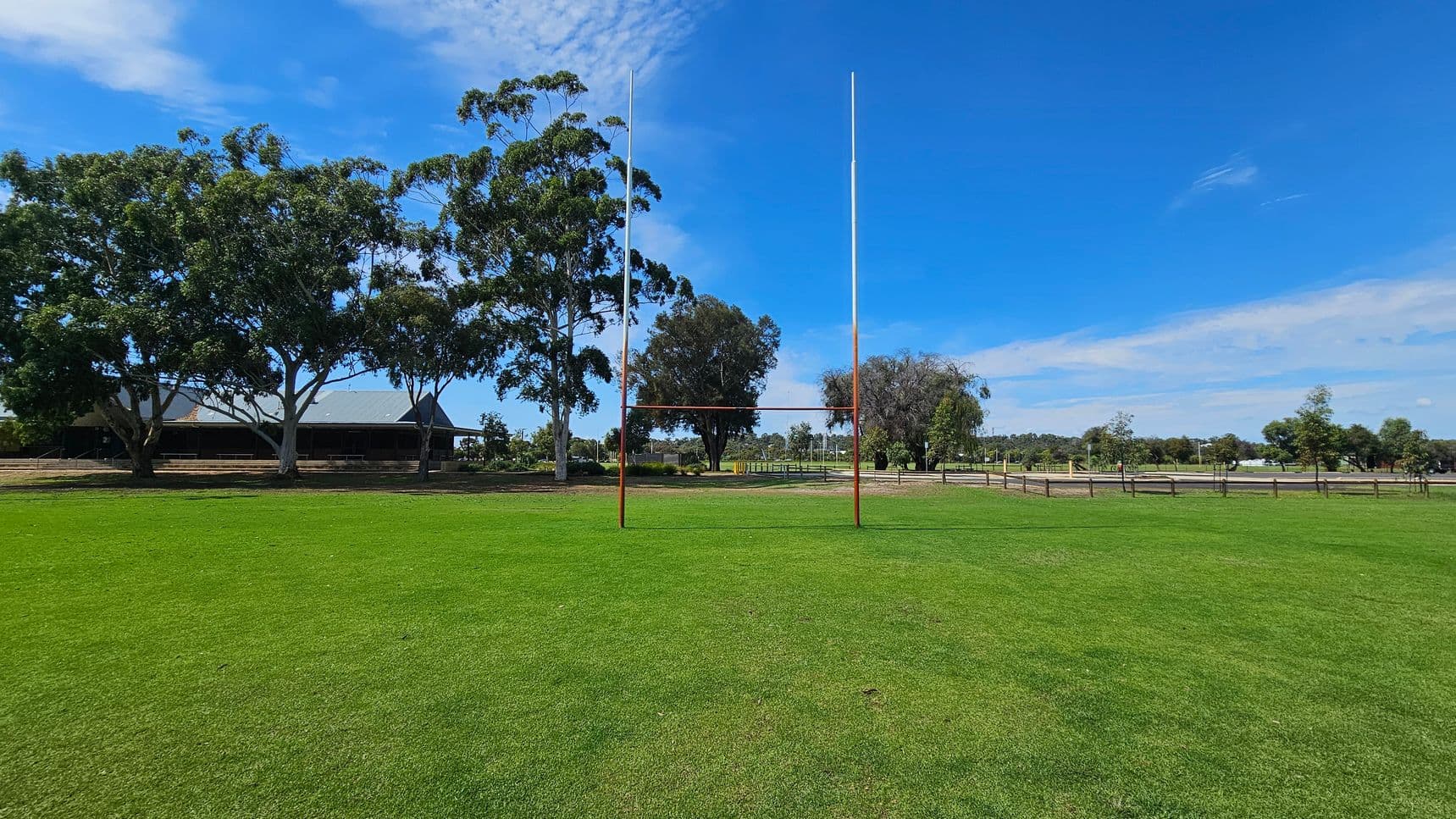 A rugby field with bright green grass is in the foreground. In the centre, there are rugby posts. To the left, large trees provide shade, and a building with a dark roof is partially visible. On the right, more trees and a wooden fence line the edge of the field. The background features a mix of trees and open space.