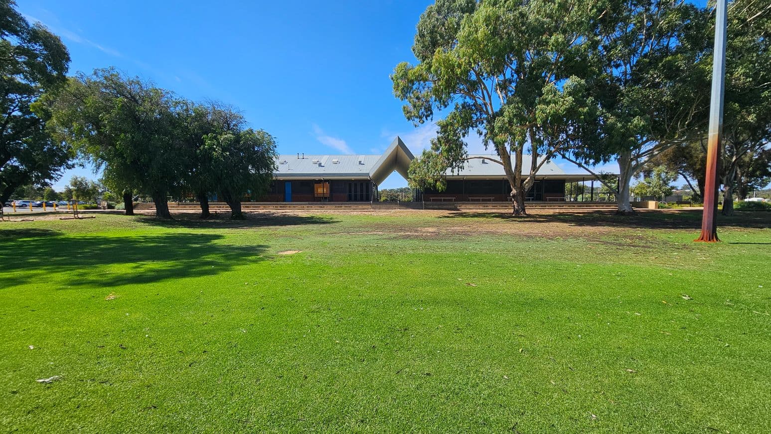 A grassy field in the foreground leads to a building with a distinctive triangular roof structure in the centre. Trees with dense foliage are on both sides, providing shade. The building has a grey roof with several vents and a blue door visible on the left side. A tall pole stands on the right side of the image. The ground near the building is a mix of grass and bare earth.