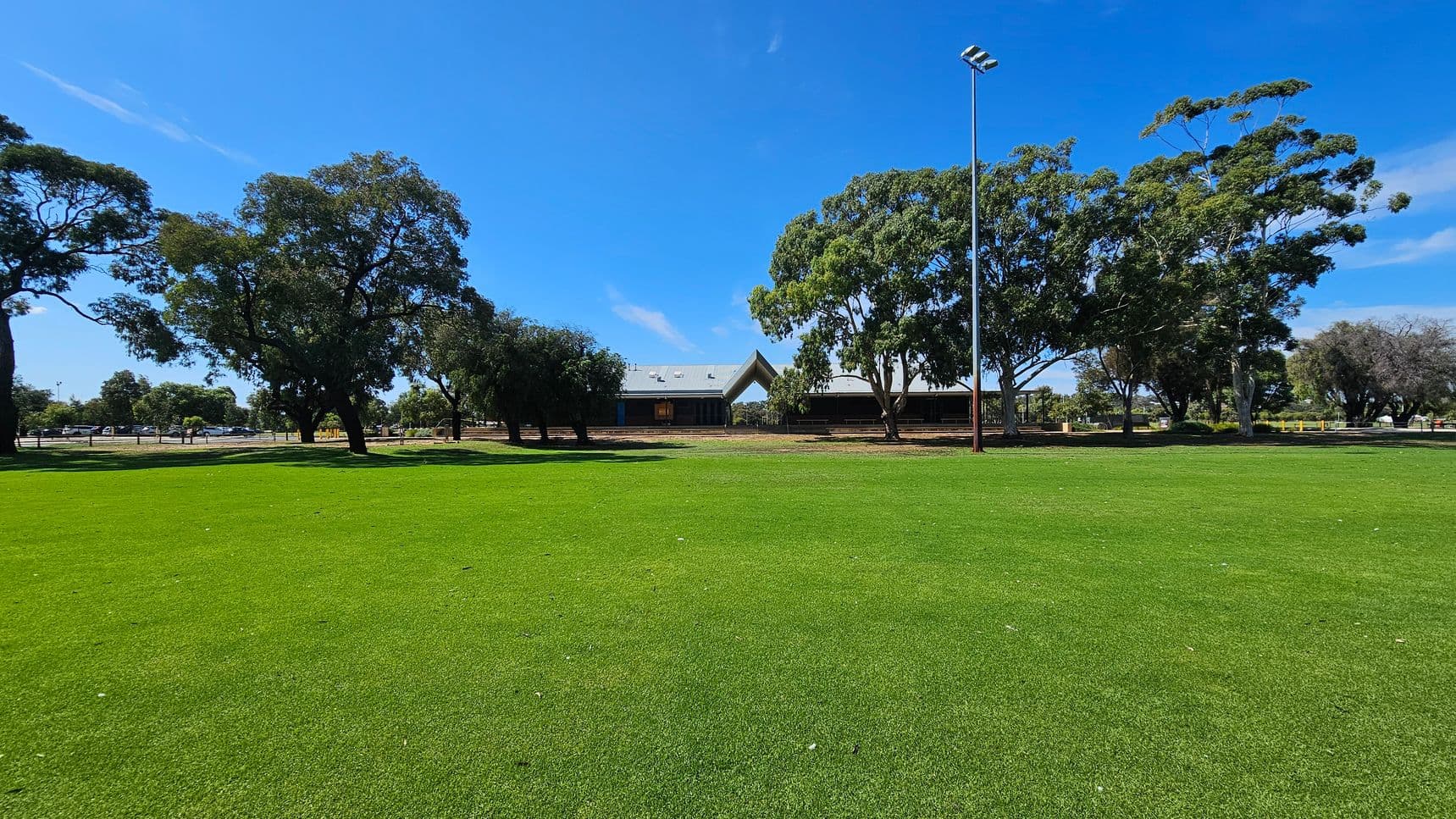 A wide expanse of green grass covers the foreground. In the background, there are several large trees with dense foliage. A building with a peaked roof is partially visible behind the trees. A tall light pole stands to the right of the building. The sky is clear and blue.